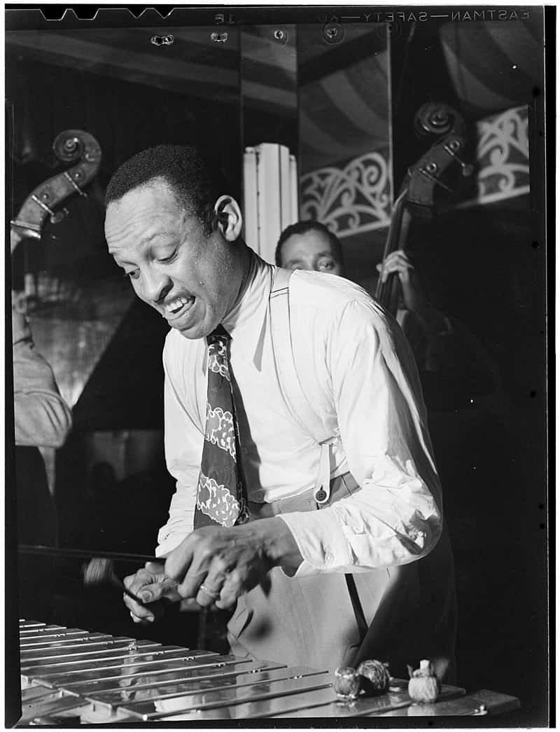 Lionel Hampton DJ 肖像照 壁紙 — 一位微笑的爵士顫音琴手，身穿白襯衫和圖案領帶，在溫暖的舞台燈光下演奏樂器，背景有一位貝斯手。