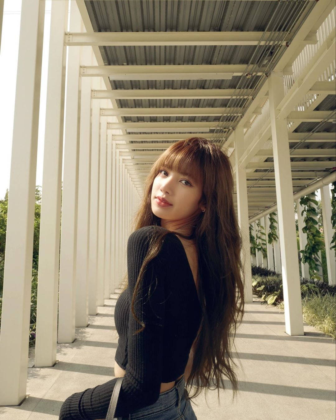 Lisa BLACKPINK泰國訪問高畫質影像 壁紙 — 一位長棕髮的年輕女性坐在白色涼棚下，涼棚在陽光燦爛的日子投下條紋狀的陰影。