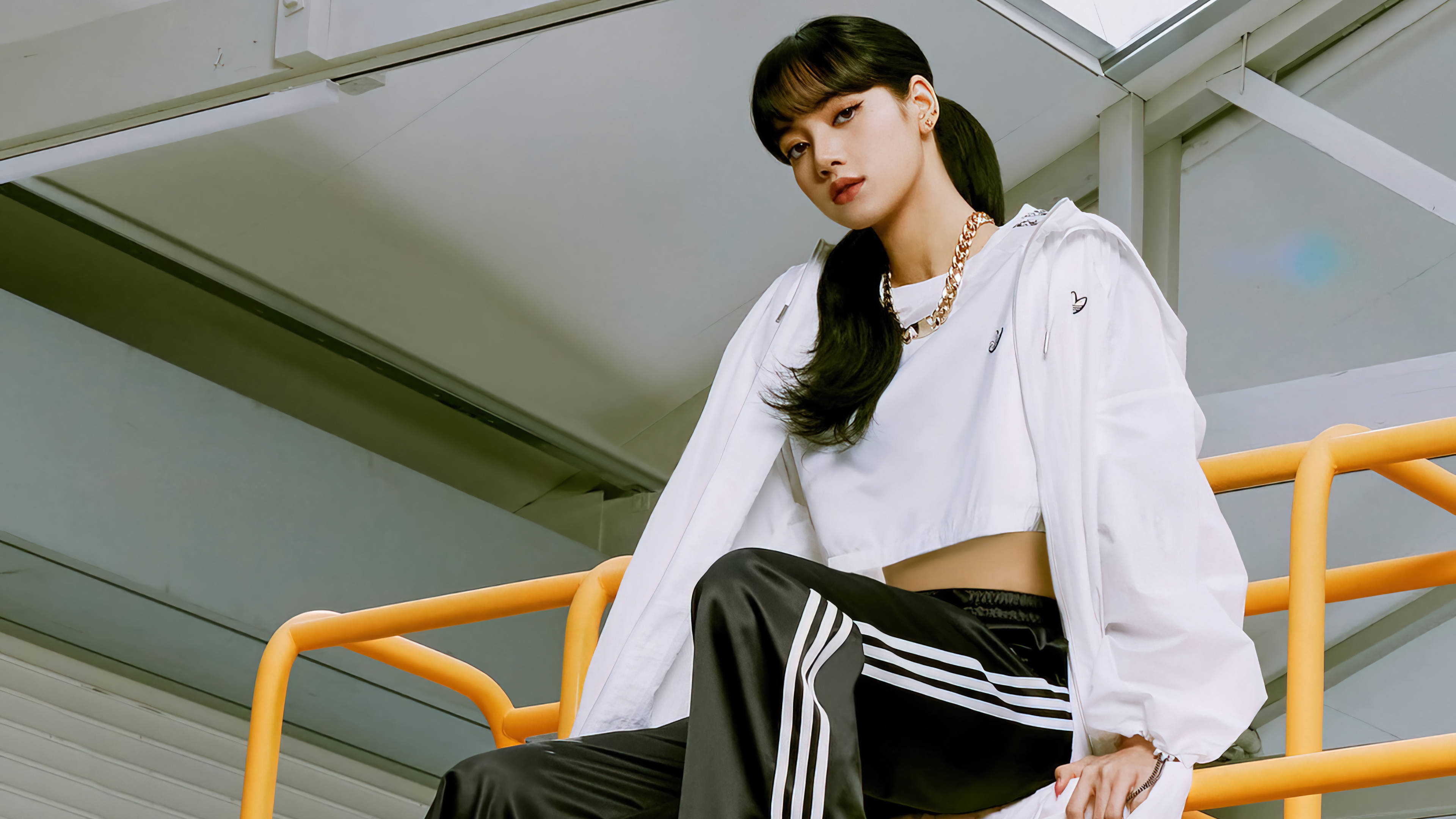 Lisa BLACKPINK與Adidas運動服廣告高清照 壁紙 — 一位身穿白色運動服的年輕女性自信地坐在工業風格燈光下的亮橙色欄杆上。