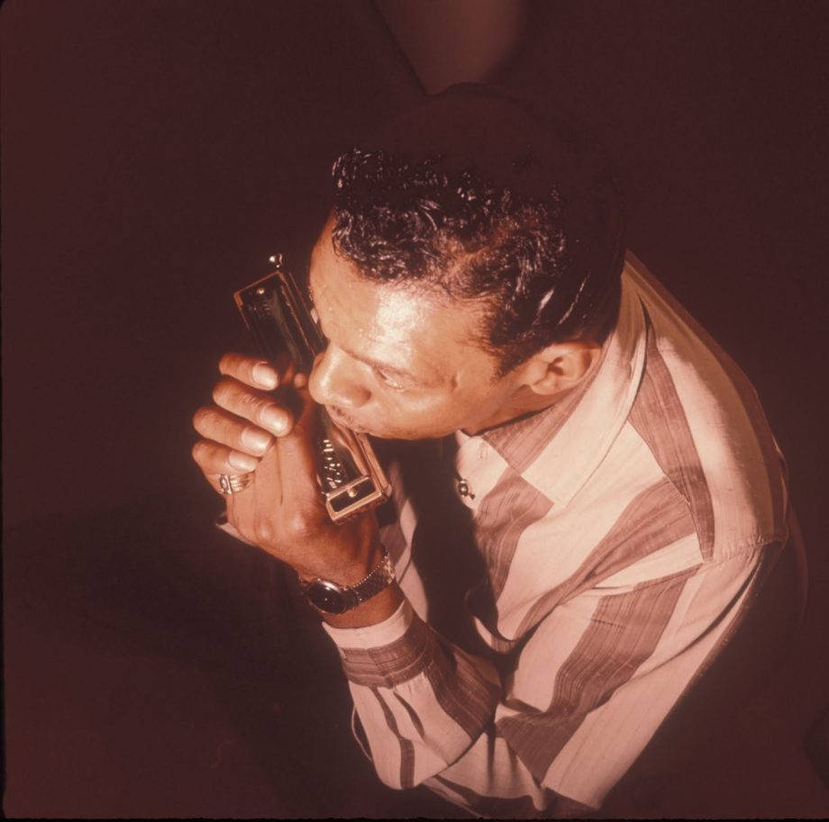 Little Walter 吹口琴空中俯拍 壁紙 — 一名身穿條紋襯衫的男子在黑暗背景下，於溫暖的聚光燈下熱情地吹奏口琴。