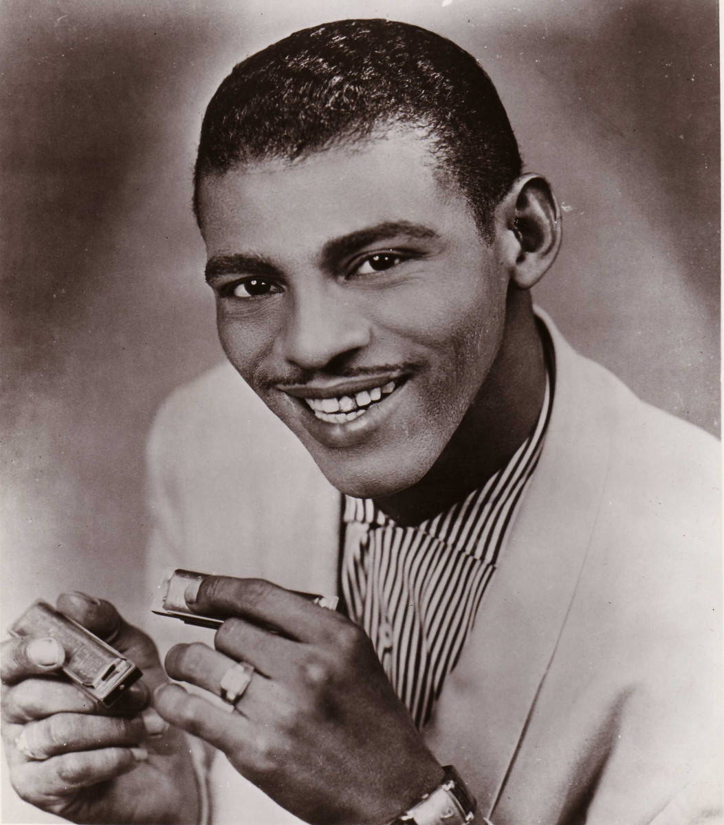 Little Walter 搞笑口琴照 壁紙 — 一位微笑的黑人男士身穿淺色西裝和條紋襯衫，在復古工作室肖像中拿著口琴和打火機。