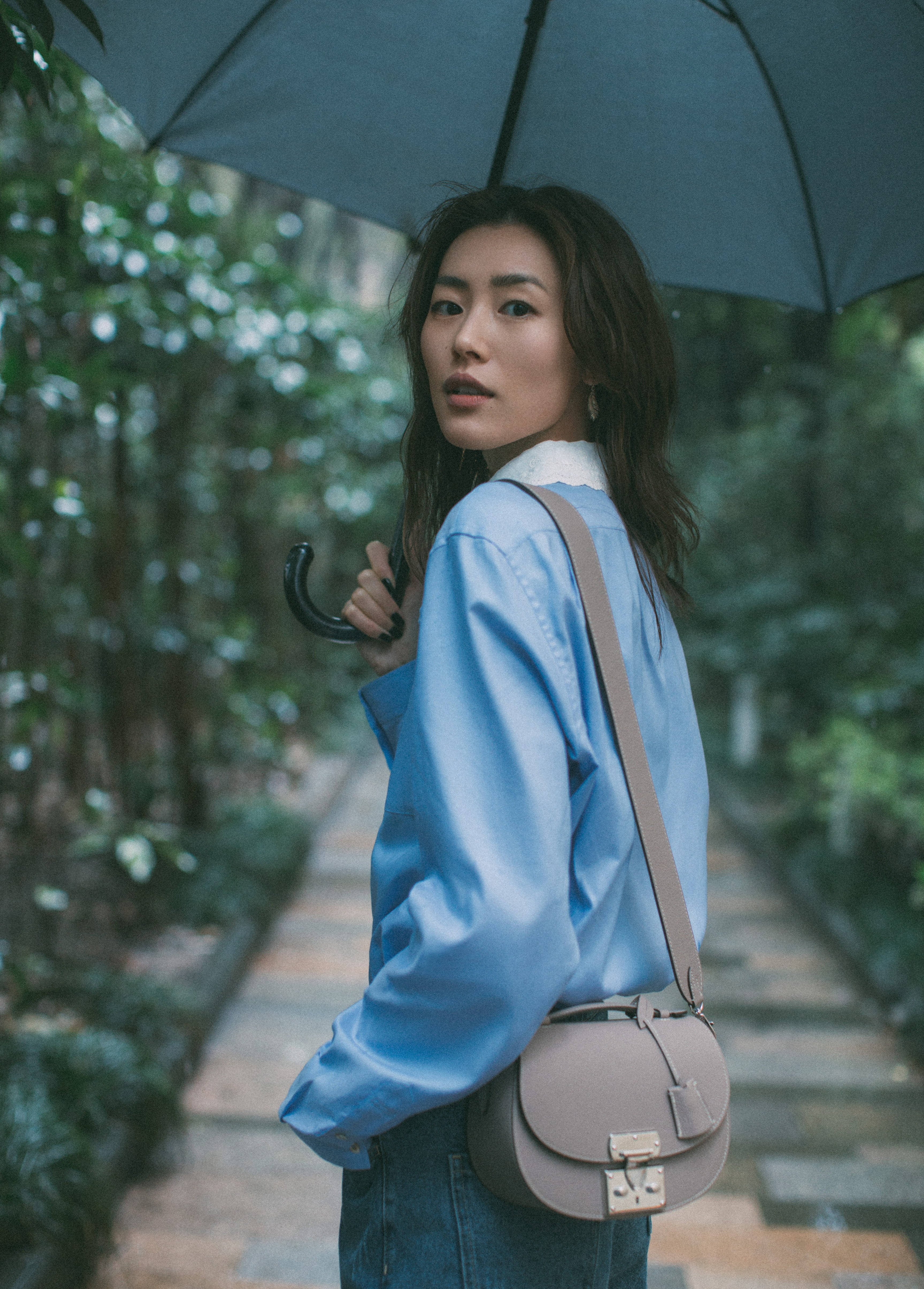 中國超模 Liu Wen 搭配 Moynat 斜背包 壁紙 — 一位身穿藍色襯衫的女子，手持深色雨傘，沿著翠綠的花園小徑行走。