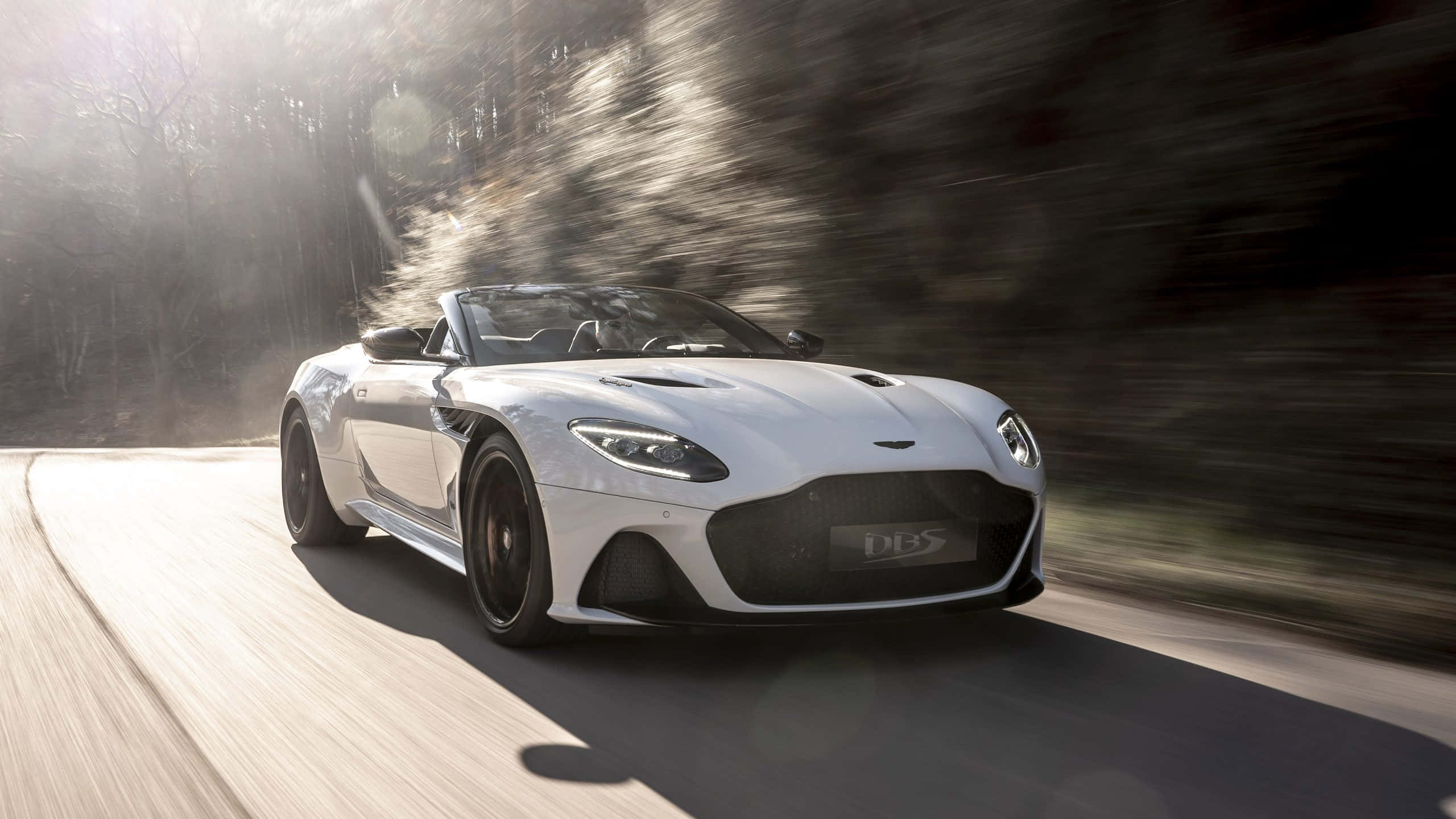 Aston Martin Dbs Superleggera 桌布 壁紙 — 一架流線型白色Aston Martin DBS Superleggera開篷跑車沿著蜿蜒的森林道路飛馳，動態模糊效果強調速度感。