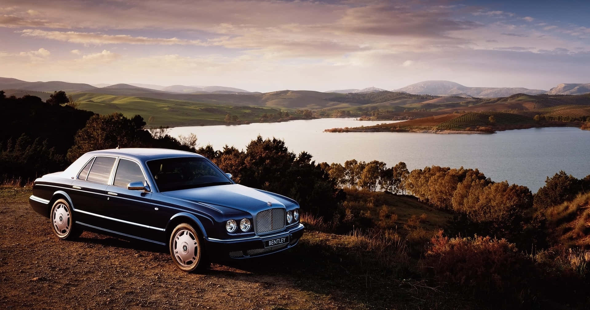 Bentley Arnage 桌布 壁紙 — 一輛流線型黑色賓利房車停泊在青草山坡上，俯瞰寧靜的湖泊和連綿起伏的山丘，天空呈現柔和的金色日落。