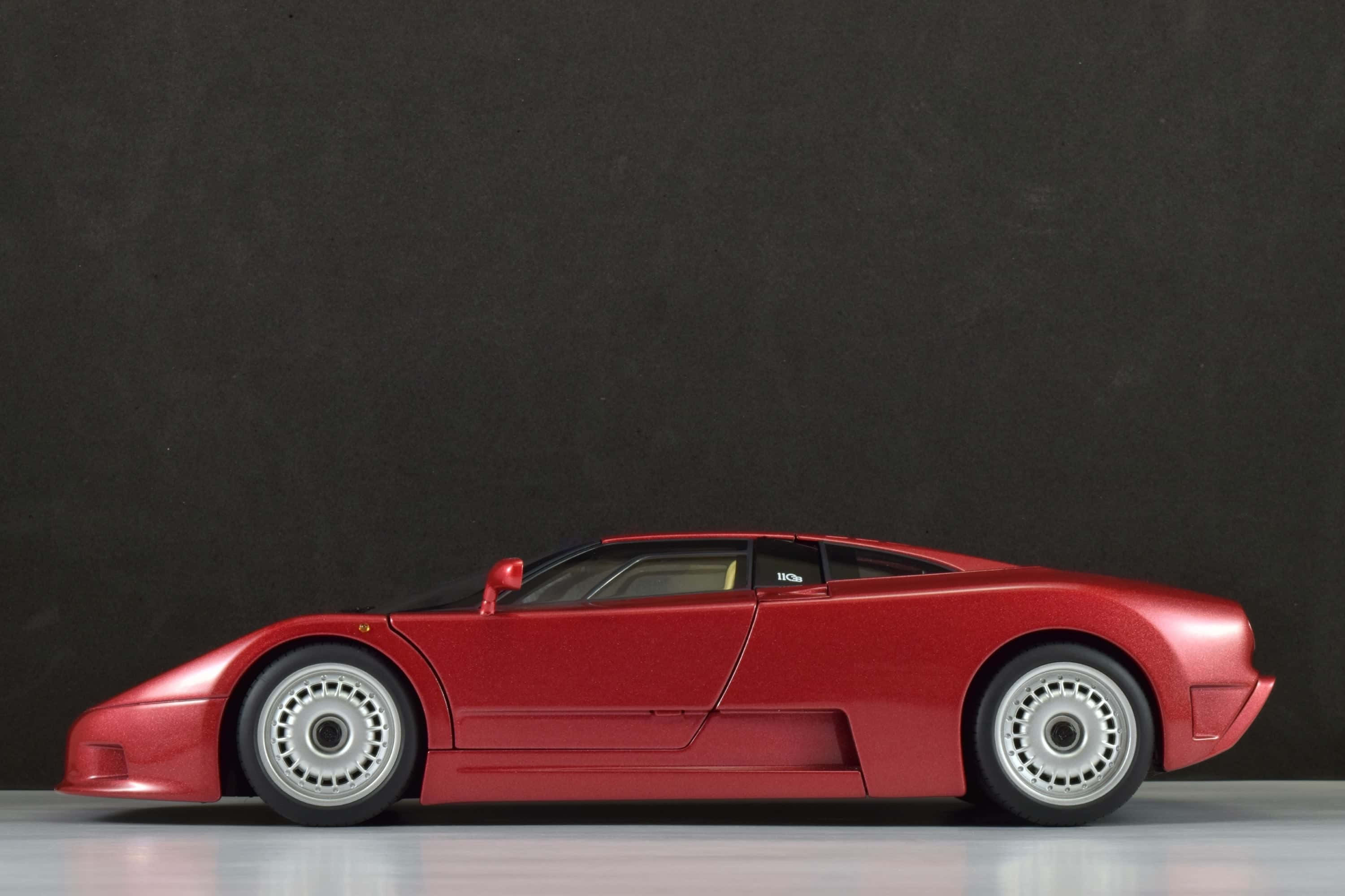 Bugatti Eb110 桌布 壁紙 — 一款流線型紅色跑車模型，配銀色輪圈，側面擺放在淺灰色地板上，背景是深色紋理牆壁。