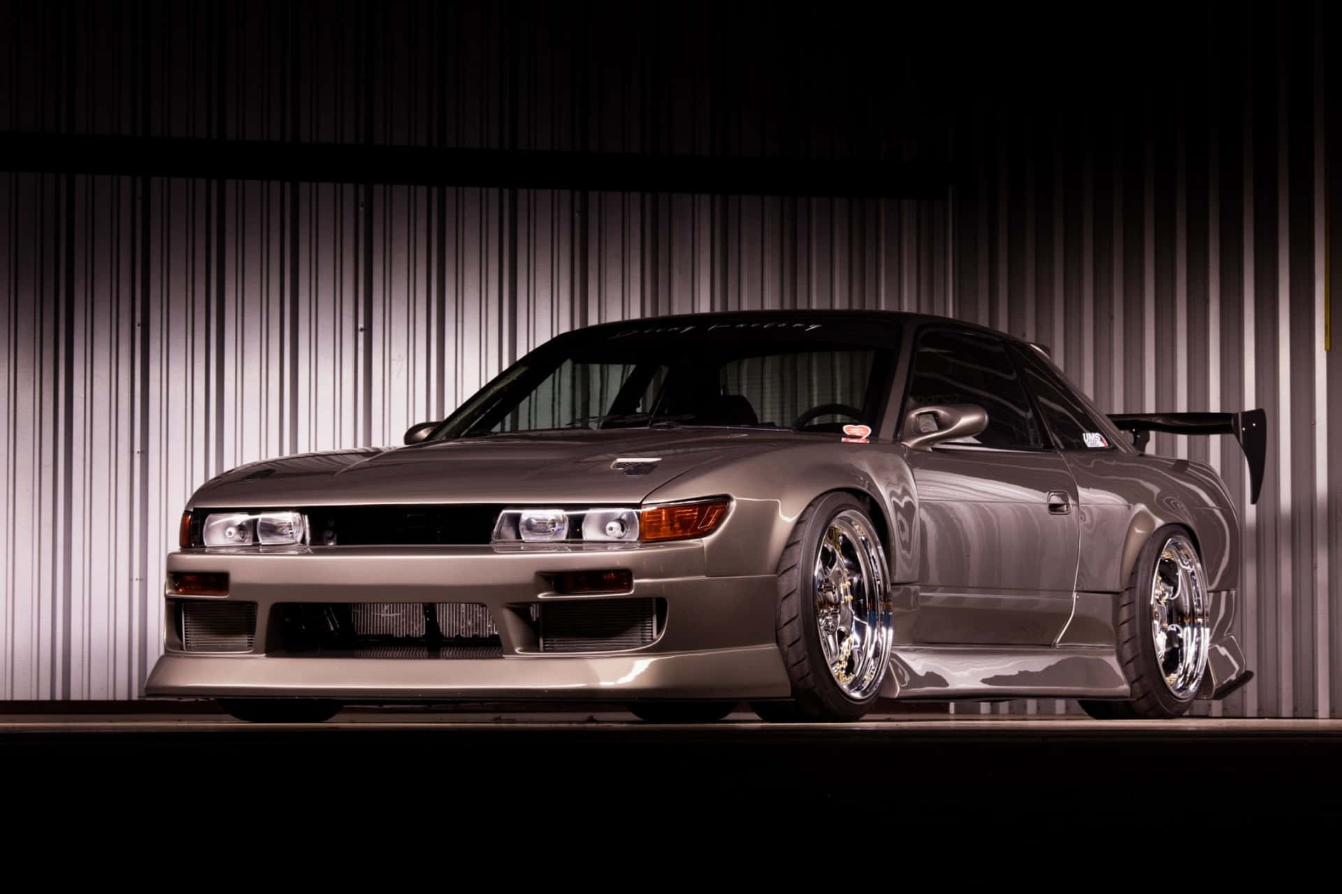  壁紙 — 一架流線型銀色日產Skyline GT-R配備定制寬體套件和鍍鉻輪圈，在波紋金屬牆前戲劇性地照亮。
