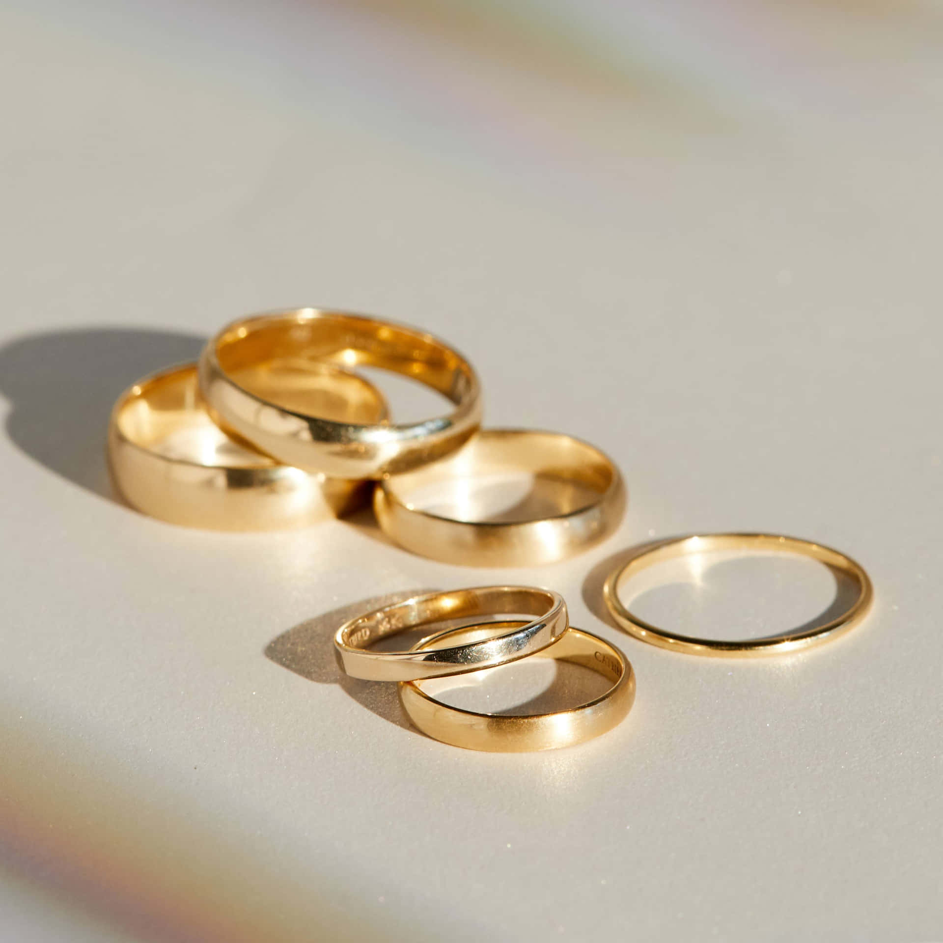 六隻金色結婚戒指排列在白色表面上，帶有柔和陰影和彩虹光。