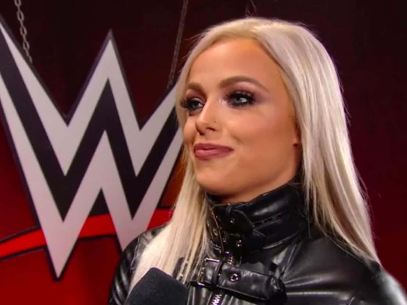 Liv Morgan WWE 專訪 壁紙 — 一位金髮女子穿著黑色皮褸，在WWE標誌前對著咪高峰微笑說話。