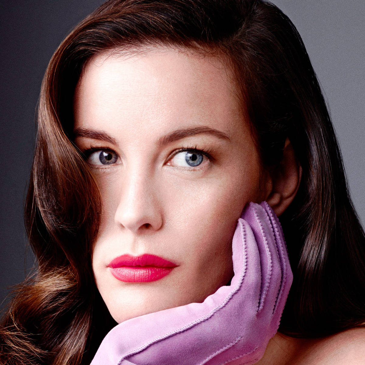 Liv Tyler 美麗模特兒 壁紙 — 一位擁有引人注目的藍色眼睛和大膽紅唇的女性，將戴著手套的手放在臉頰附近，背景是柔和的灰色。