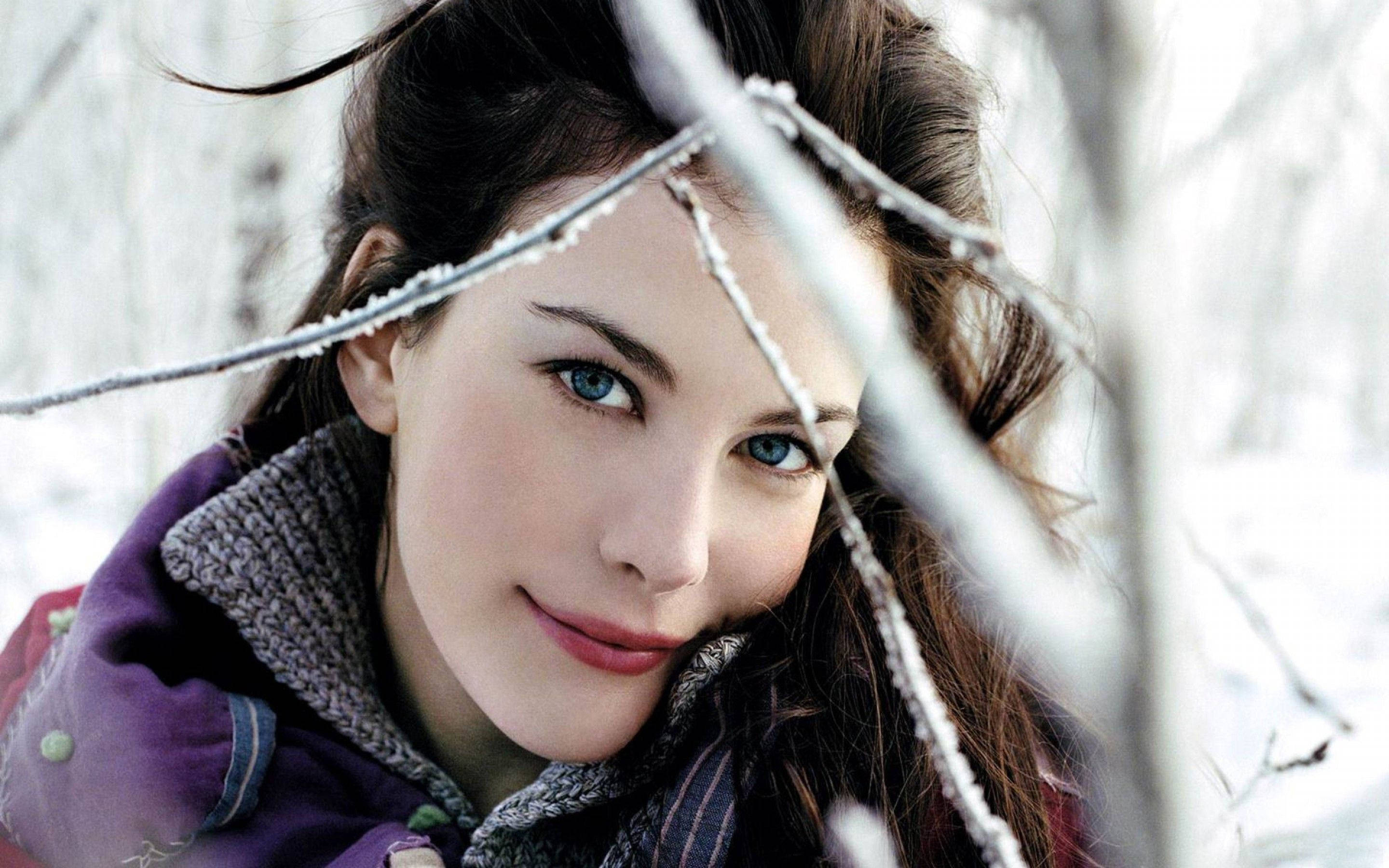 Liv Tyler 冬季時尚模特兒 壁紙 — 一位擁有引人注目的藍眼睛的女性，在白雪皚皚的冬季森林中，透過結霜的樹枝溫柔地微笑。
