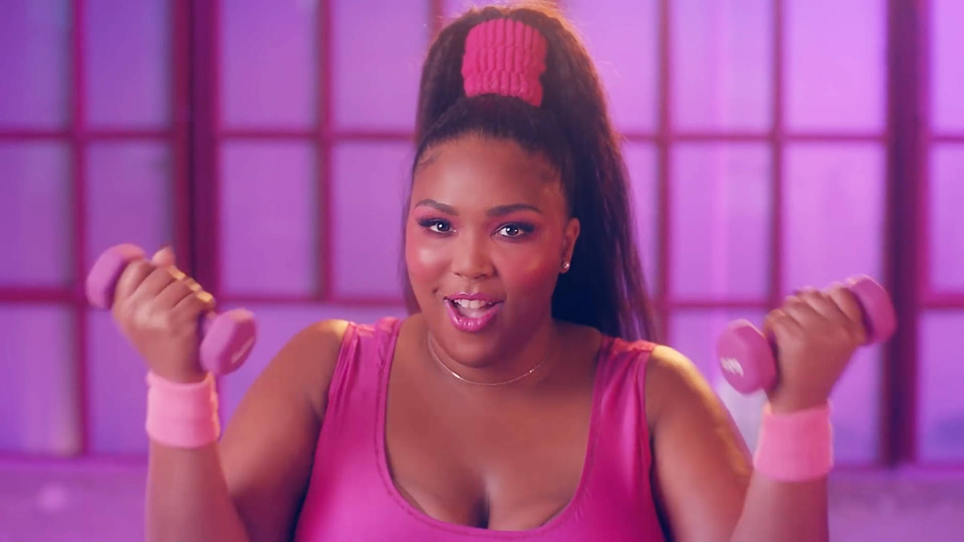 Lizzo《Juice》音樂錄影帶劇照 壁紙 — 一位紮著高馬尾、佩戴粉色配飾的女性，在紫色鑲板背景前微笑著舉起粉色啞鈴。