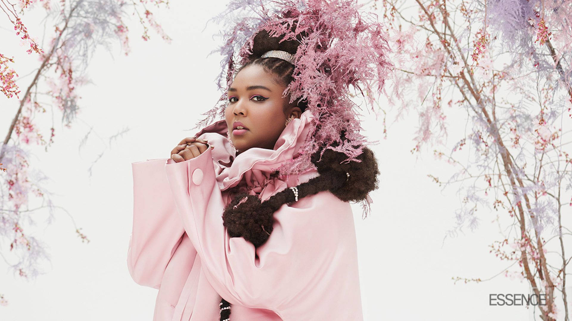 Lizzo 為 Soul Glo 拍攝數位封面 壁紙 — 一位身穿粉紅色外套和羽毛頭飾的女性，站在盛開的粉紅色和紫色樹木下，背景是柔和的白色天空。