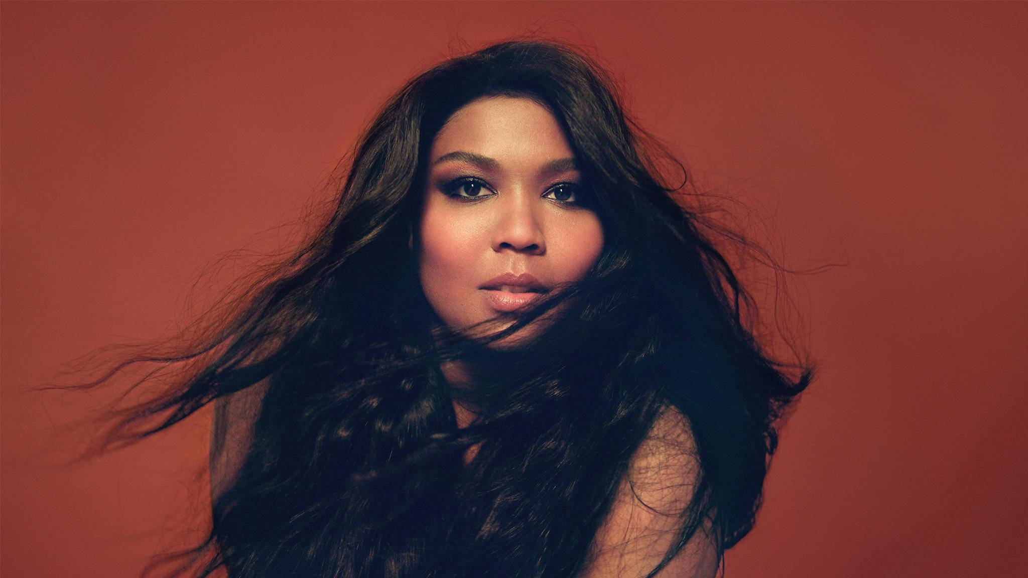 Lizzo 紅棕色背景照 壁紙 — 一位長長深色頭髮的女性，頭髮在溫暖的陶土色背景前吹過她的臉龐。