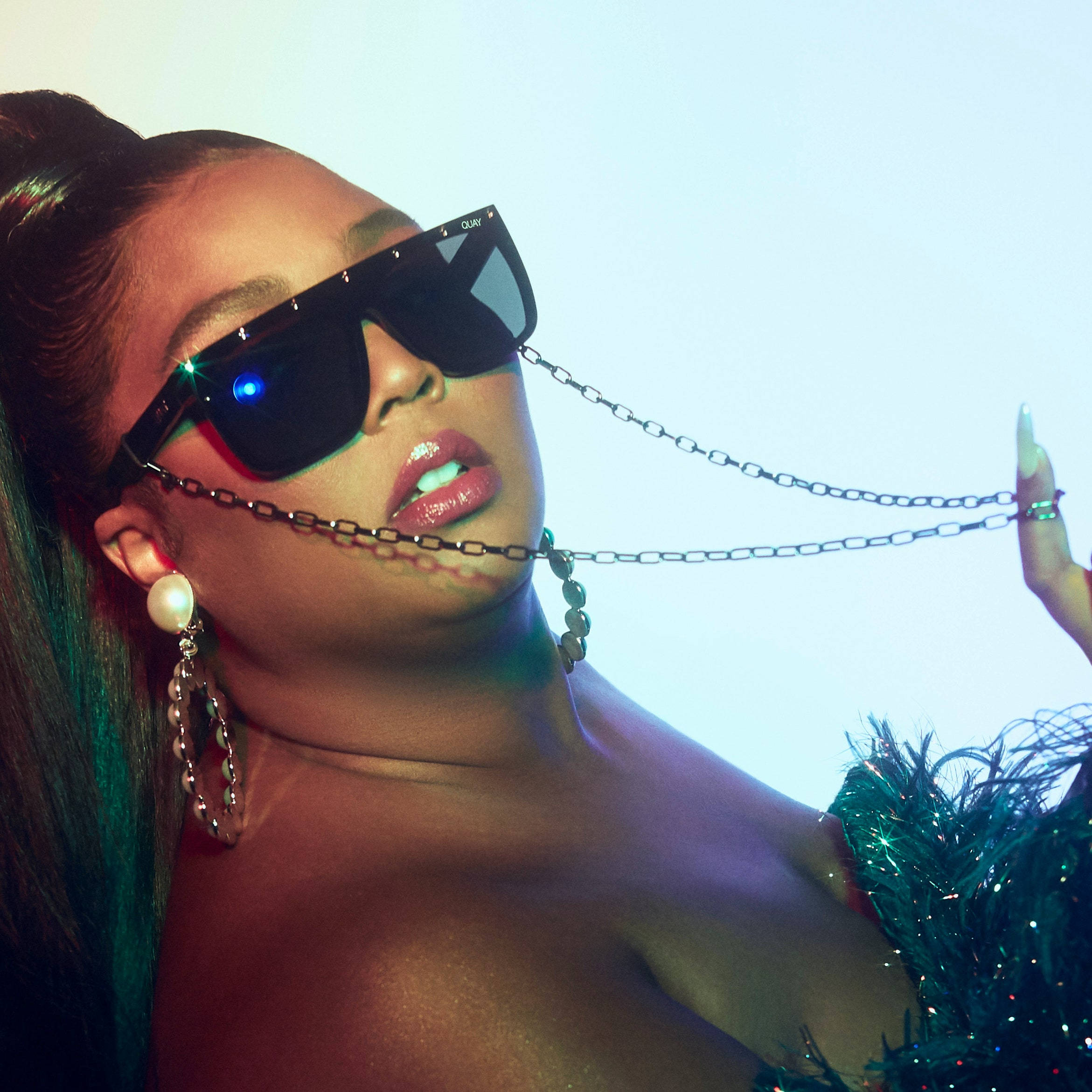 Lizzo 高級黑墨鏡 壁紙 — 一位時尚女性戴著大膽的太陽眼鏡和鏈條飾品，在發光的藍色背景前擺姿勢。