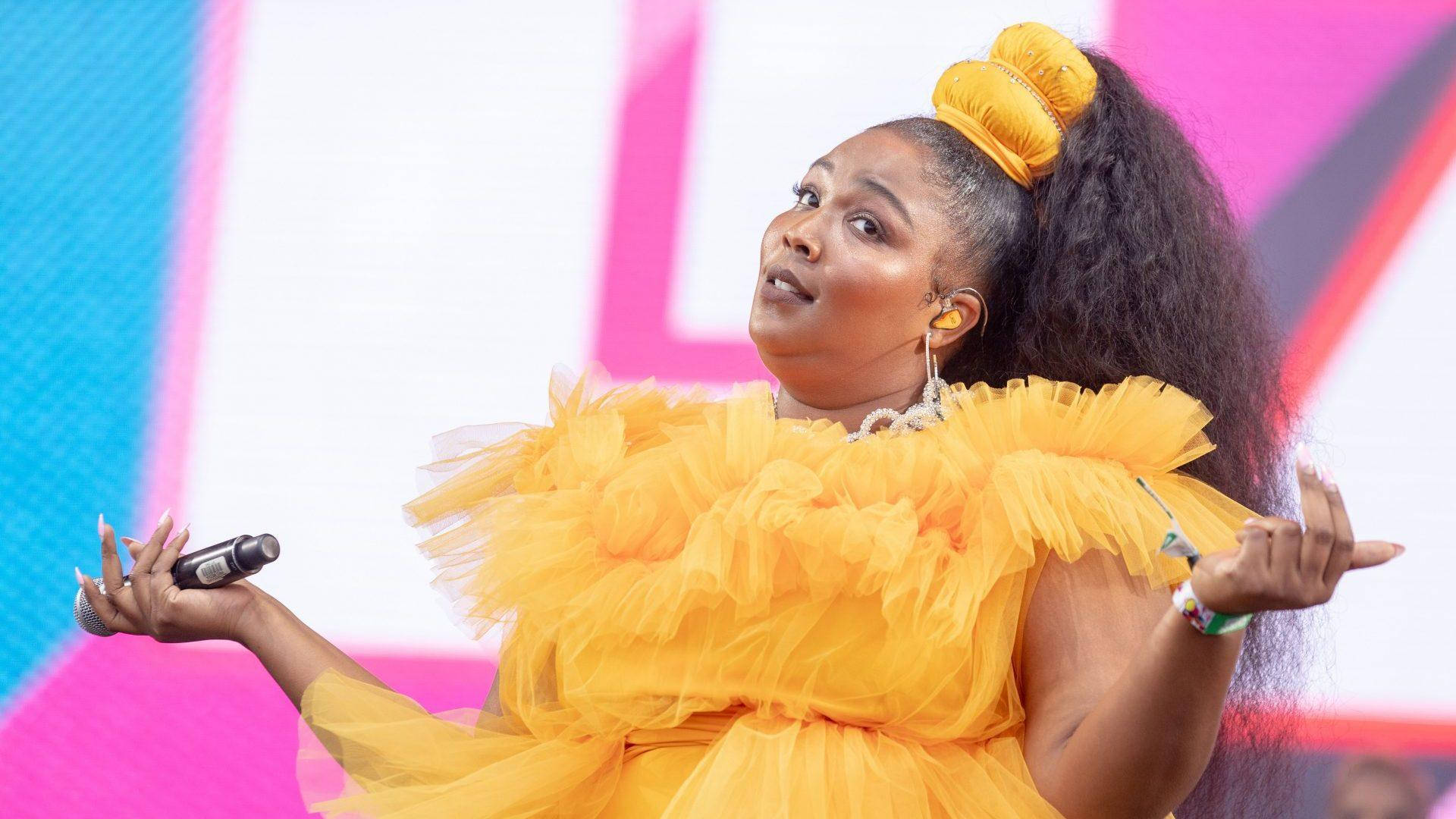Lizzo 黃色蕾絲服裝演出 壁紙 — 一位充滿活力的表演者身穿荷葉邊黃色連衣裙，在色彩繽紛的舞台上熱情歌唱，身後是醒目的粉紅色和藍色幾何燈光。