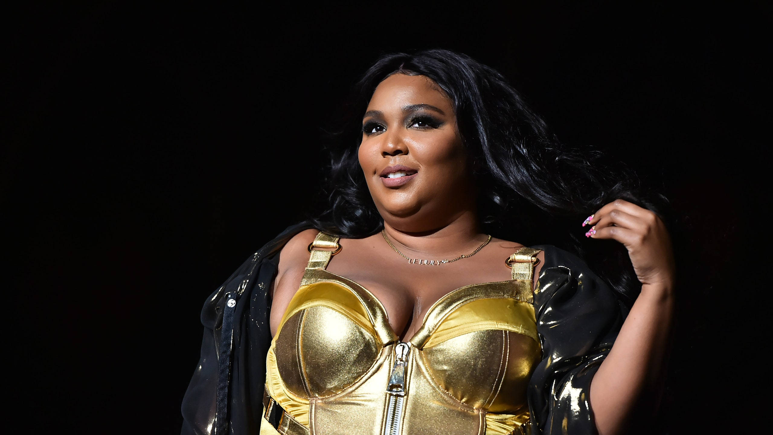 Lizzo 黑金皮革造型 壁紙 — 一位身穿閃亮金色緊身胸衣和黑色外套的女性，在深色背景下自信地擺姿勢，頭髮飄逸。