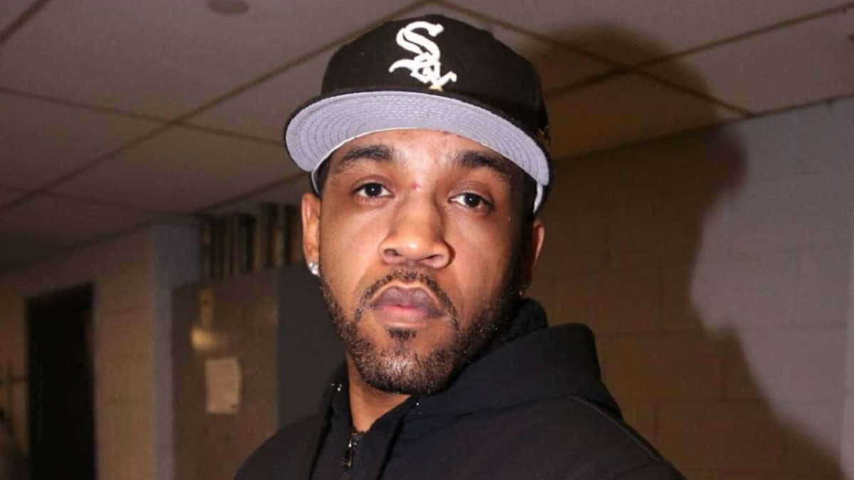 Lloyd Banks 黑色連帽衫與白邊棒球帽 壁紙 — 一位留鬍鬚、戴黑色棒球帽的男子在光線昏暗的室內走廊直視鏡頭。