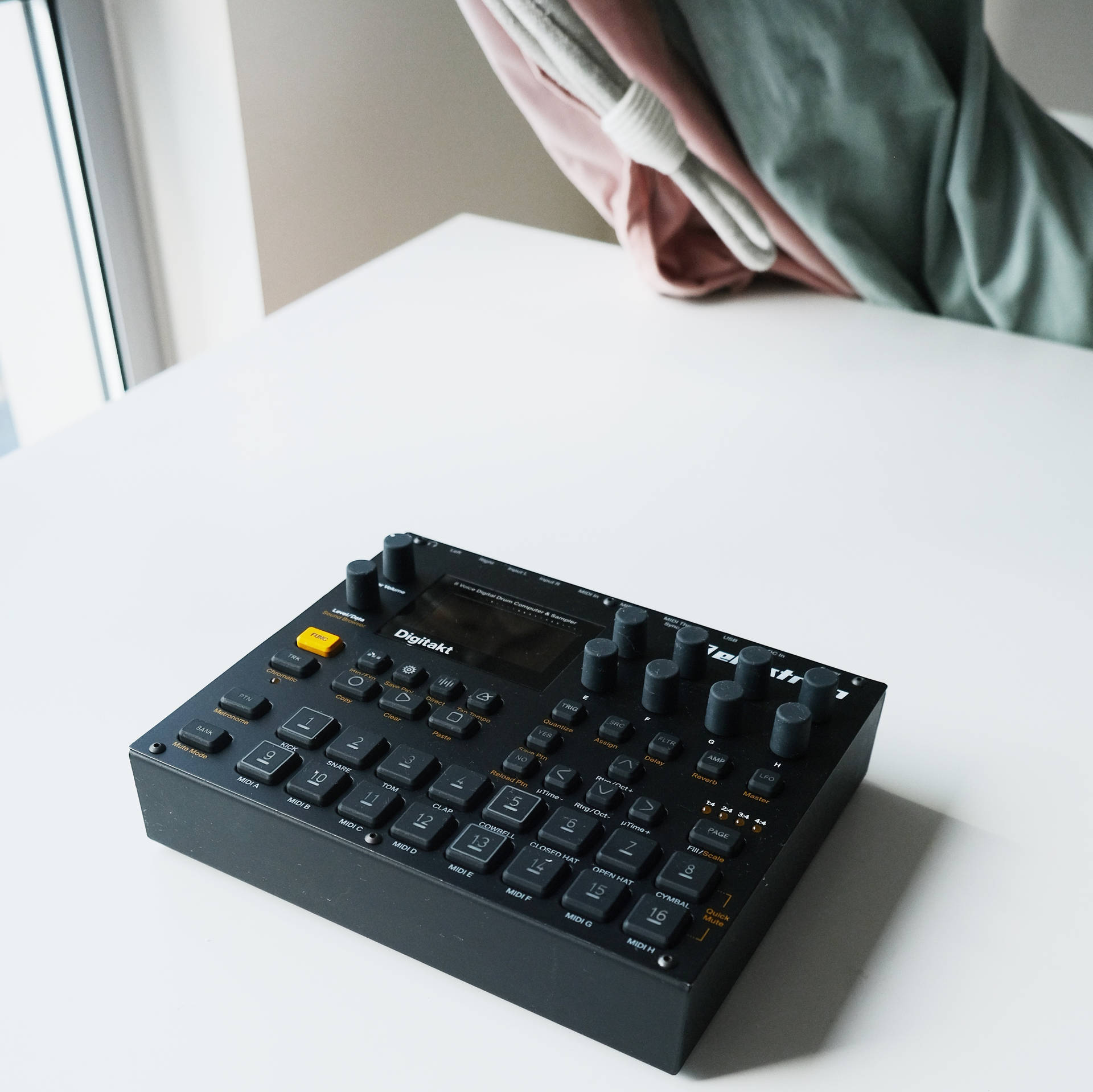 窗邊白色桌上的黑色 Digitakt 音樂序列器，帶有垂掛布料。