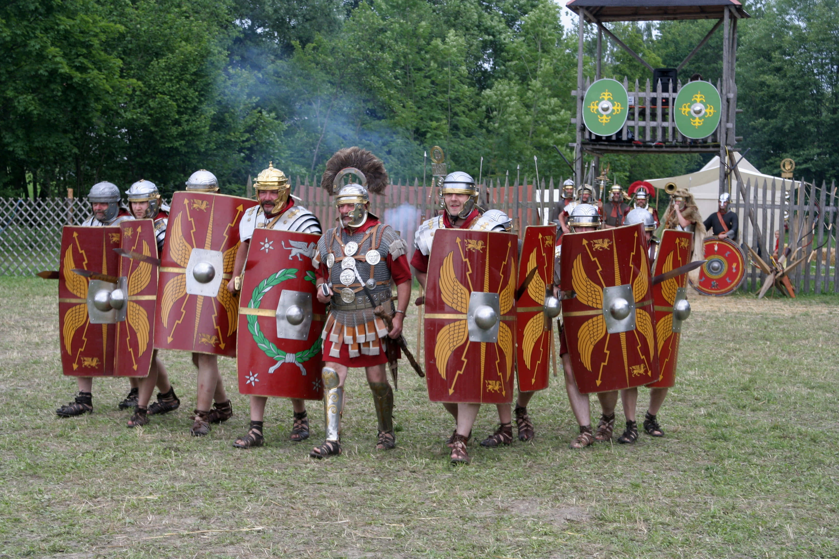 Roman 桌布 壁紙 — 羅馬軍團士兵身穿精細盔甲和紅色盾牌，在歷史重演活動中於木造堡壘附近的草地上列隊。