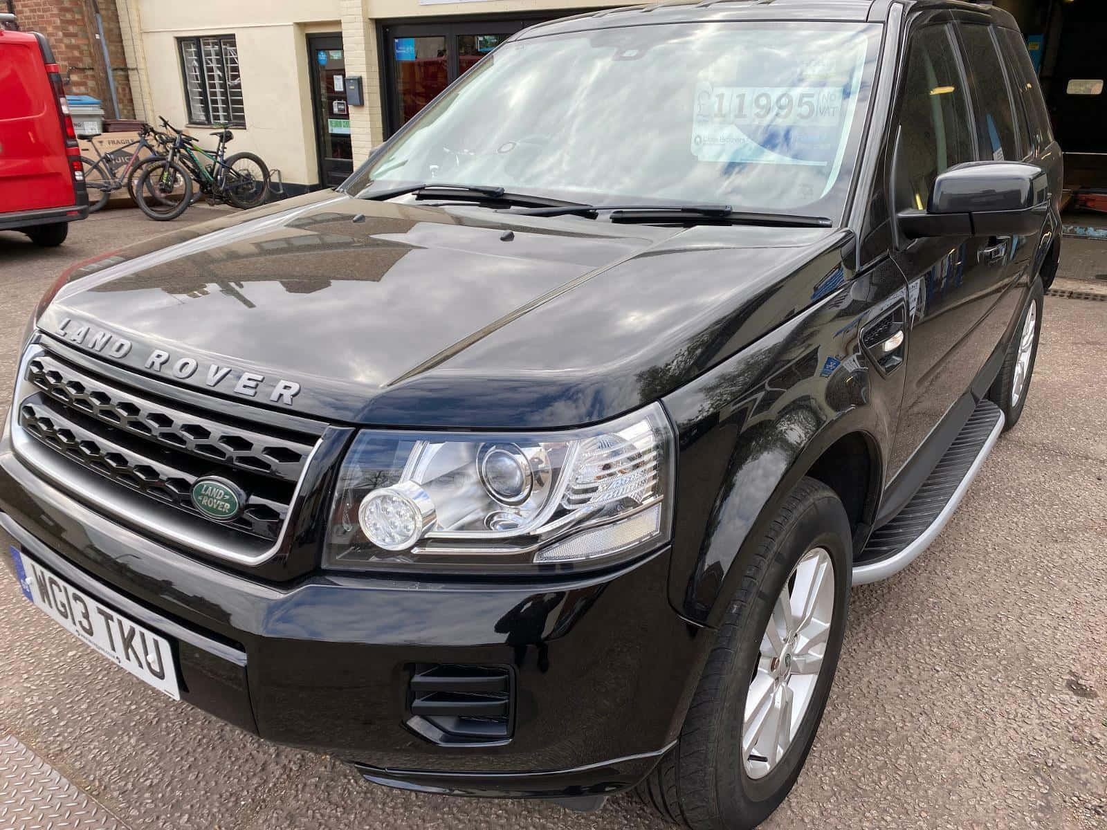 Land Rover Freelander 桌布 壁紙 — 一架亮黑色嘅Land Rover Freelander泊喺鋪砌嘅停車場，背景有單車同紅色貨車。