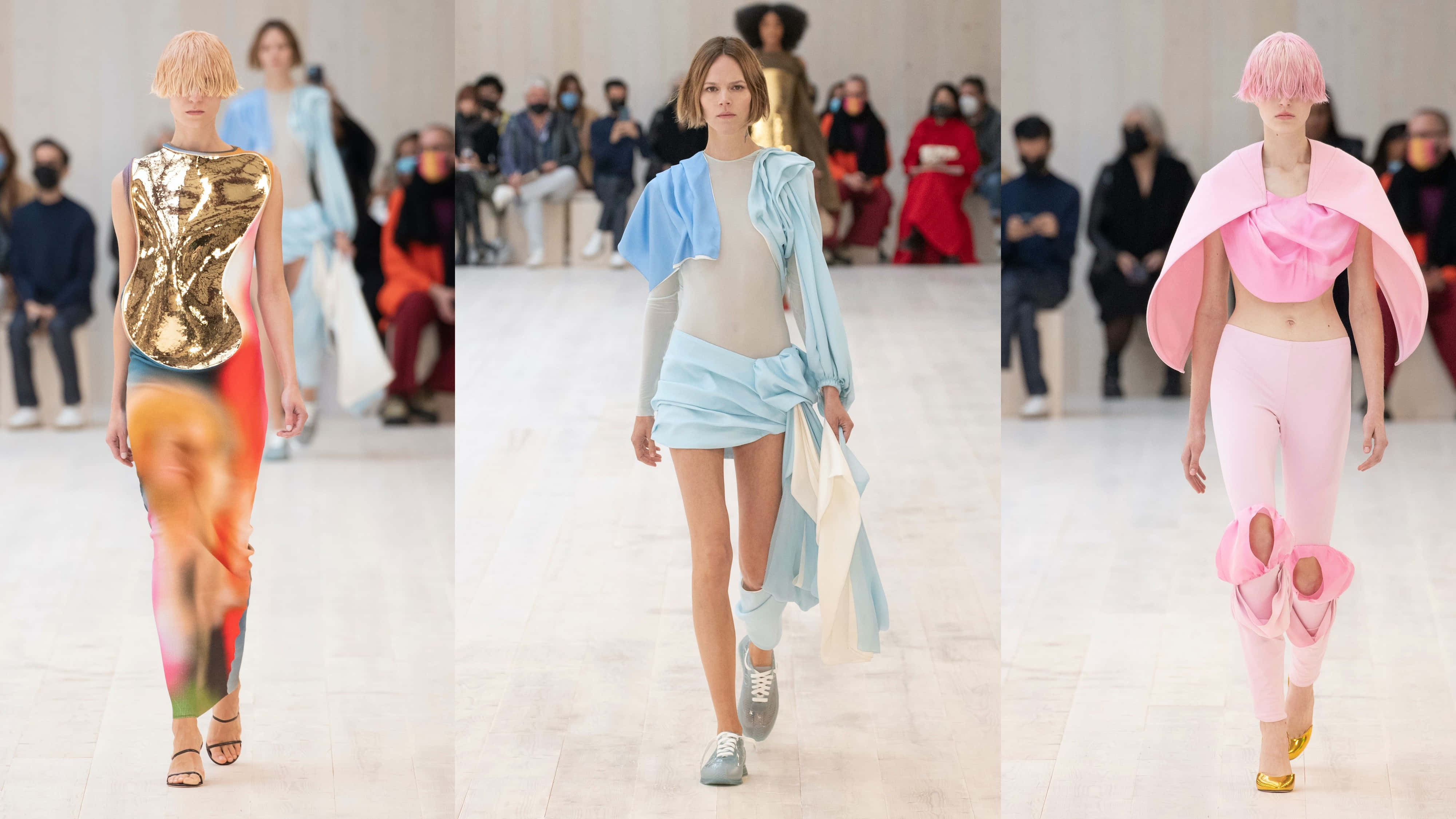 Loewe 2022春夏女裝系列 壁紙 — 三位模特兒在明亮的時裝天橋上走秀，展示前衛服裝，包括金屬金色、粉藍色和粉紅色荷葉邊套裝。