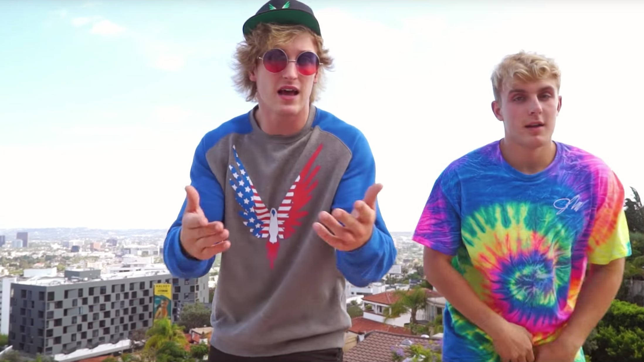 Logan Paul 拳擊訓練日常 壁紙 — 兩名身穿彩色紮染襯衫和太陽眼鏡的年輕男子在明亮的日光下，在城市天際線上充滿活力地擺姿勢。