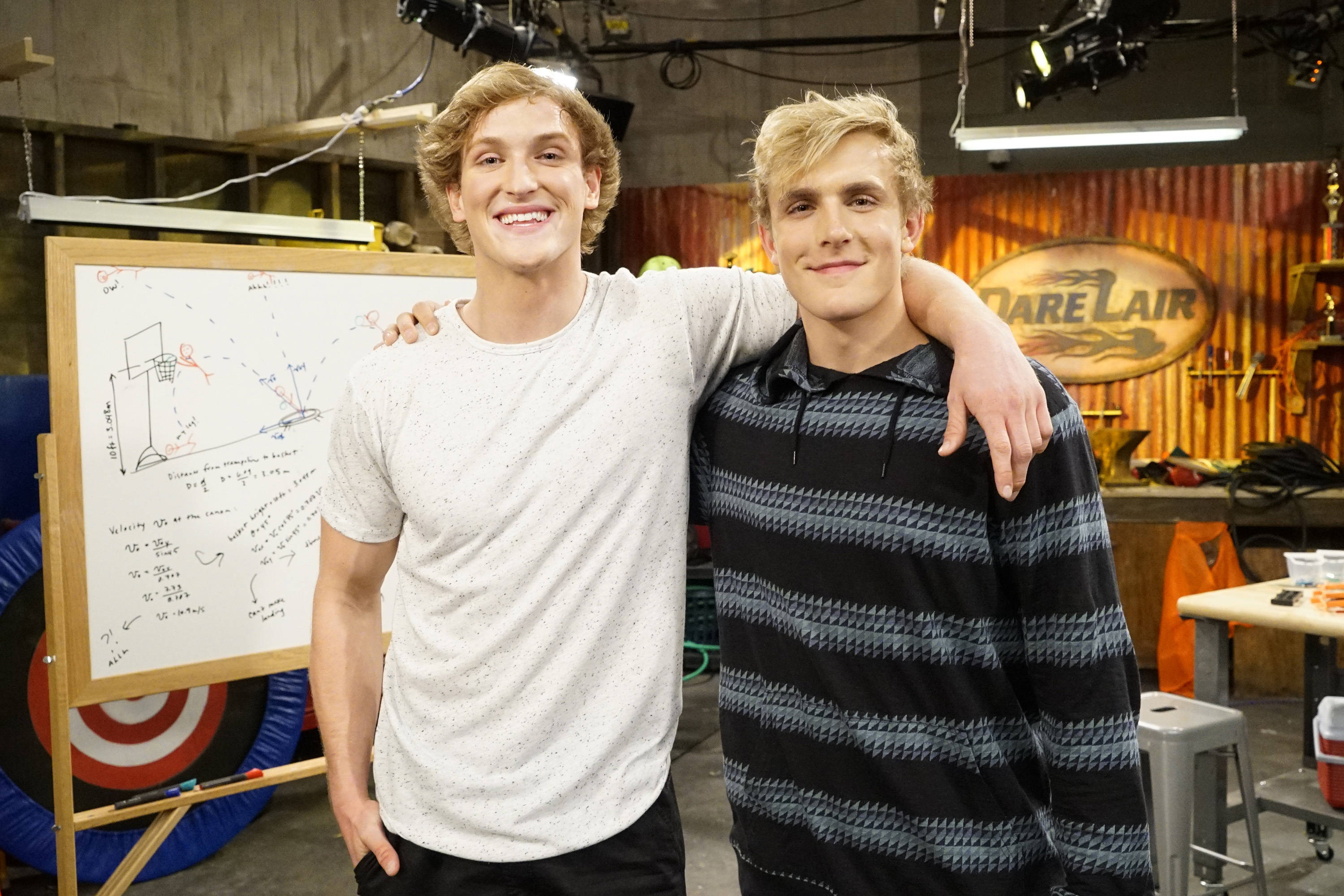 Logan Paul 出現在迪士尼喜劇劇集 壁紙 — 兩個微笑的年輕男子在雜亂的工作室中手挽手擺姿勢，身後有「Dare Lair」標誌和籃球策略板。