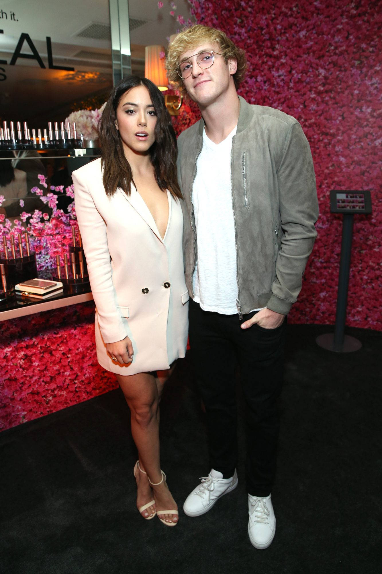 Logan Paul 與 Chloe Bennet 化妝品活動 壁紙 — 一名男子和一名女子在一個華麗的活動中，在充滿活力的粉紅色花牆前一起擺姿勢。