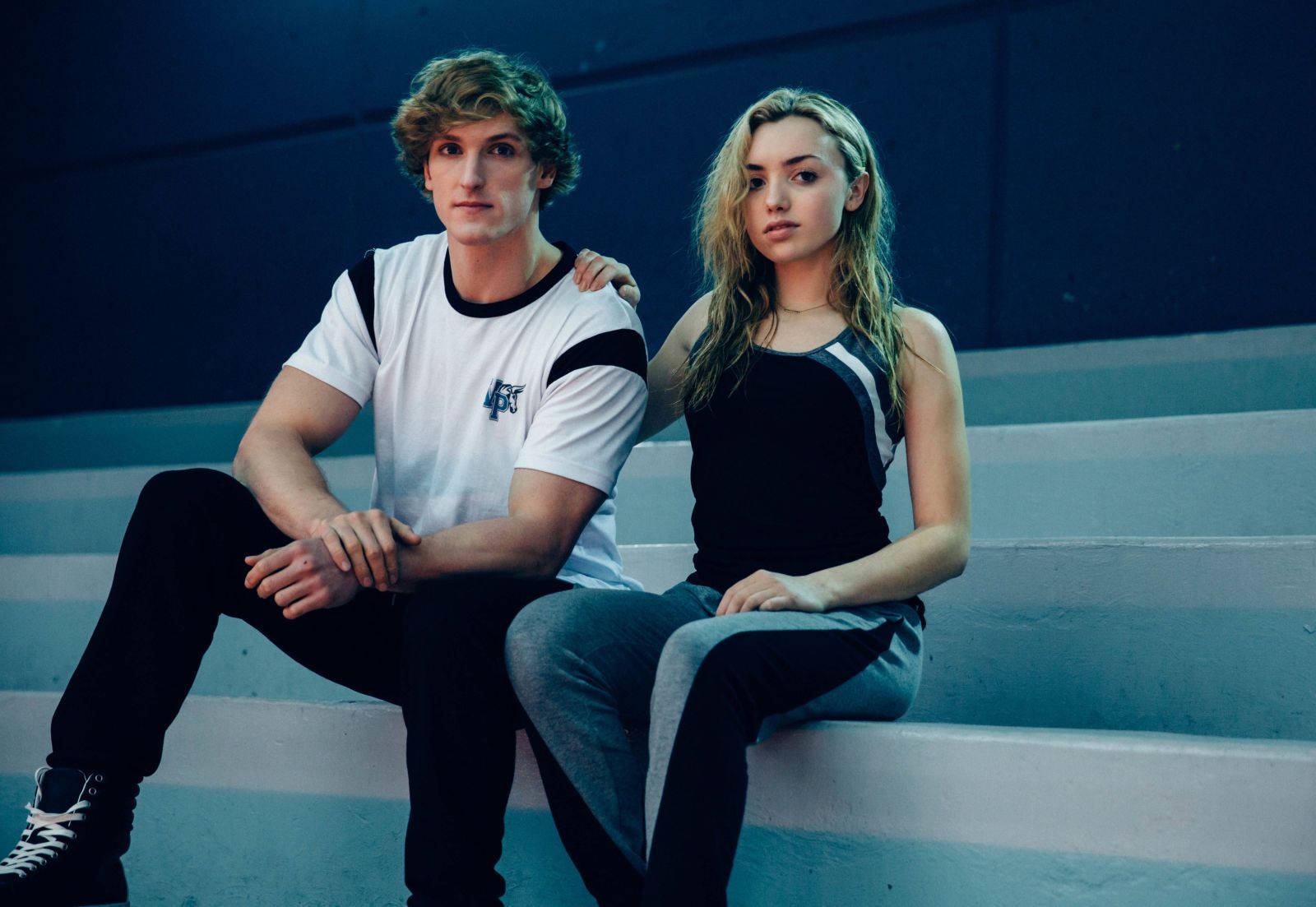 Logan Paul 與 Peyton List 合照 壁紙 — 一名年輕男子和女子在冷藍色燈光下緊密地坐在混凝土台階上，兩人都直視鏡頭，表情嚴肅。