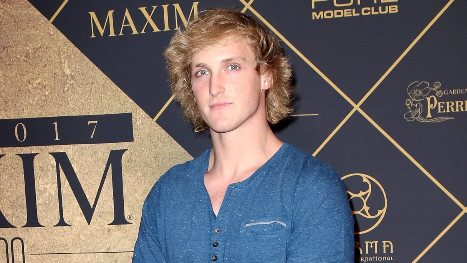 Logan Paul 身穿藍色亨利衫 壁紙 — 一位年輕男子，波浪形金髮，穿著藍色亨利衫，背景為品牌活動背景。