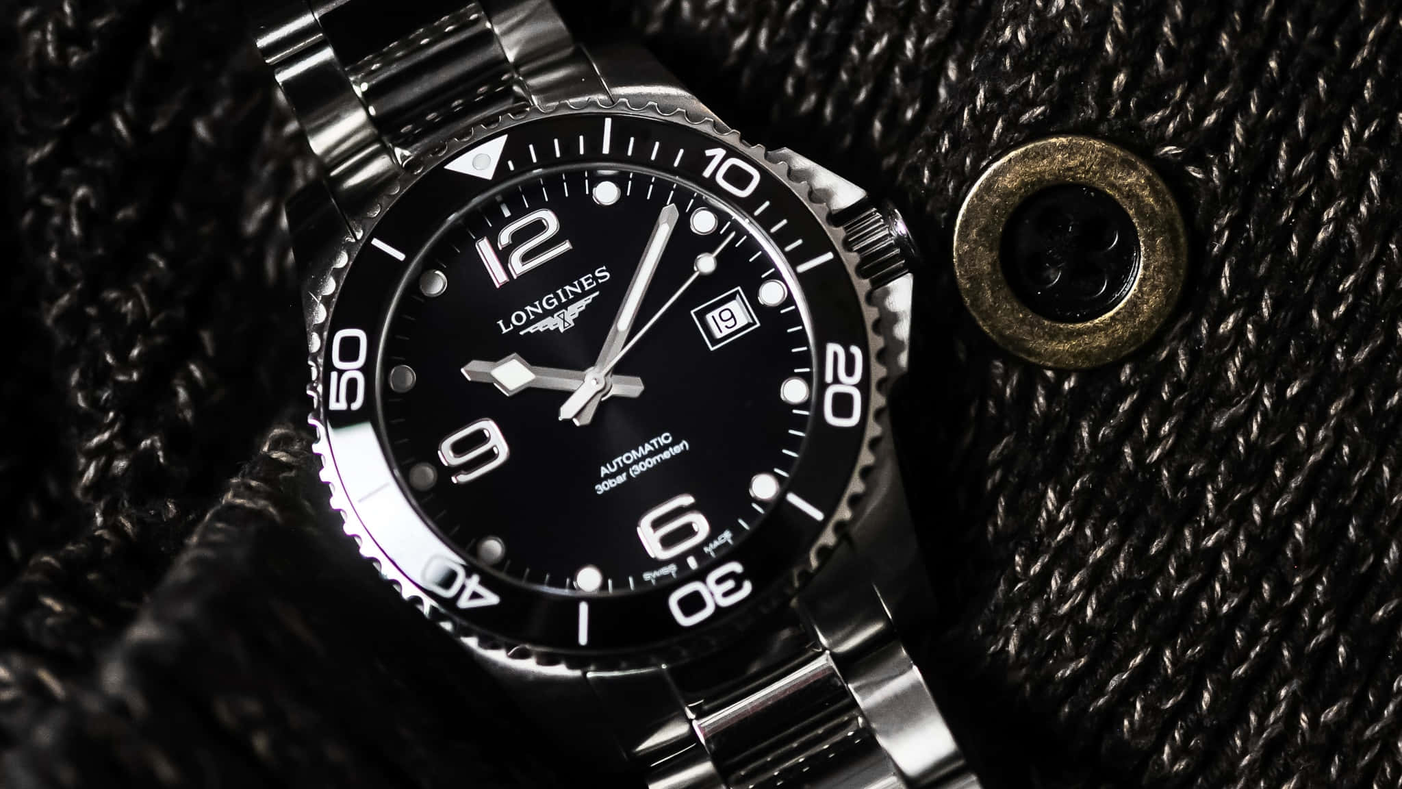 Longines HydroConquest腕錶置於織物背景上 壁紙 — 一隻時尚的銀色Longines手錶，黑色錶盤，放在有紋理的黑色布料上，旁邊有一個黃銅鈕扣。