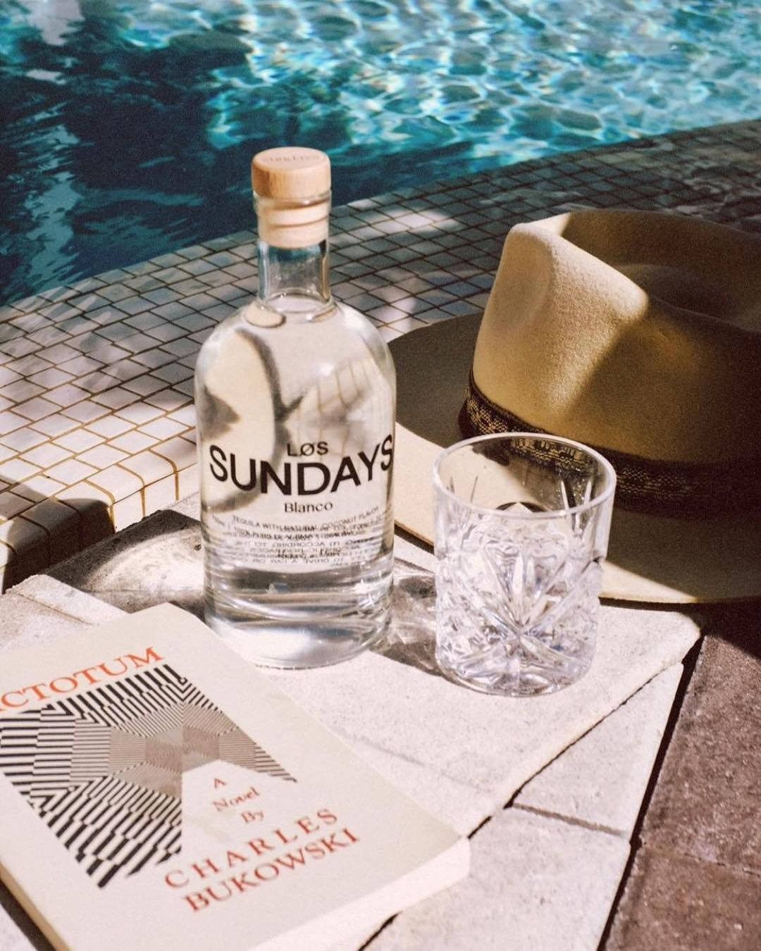 Los Sundays泳池畔 壁紙 — 一瓶 Los Sundays Blanco 龍舌蘭酒、玻璃杯和軟呢帽放在陽光普照的泳池邊瓷磚上，旁邊有一本書。