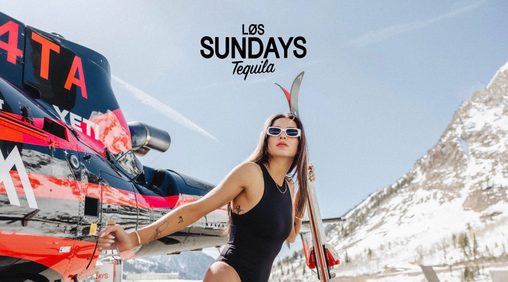 Los Sundays龍舌蘭酒廣告 壁紙 — 一名身穿黑色泳衣的女子在晴朗藍天下，以積雪山脈為背景，手持滑雪板站在紅黑相間的直升機旁。