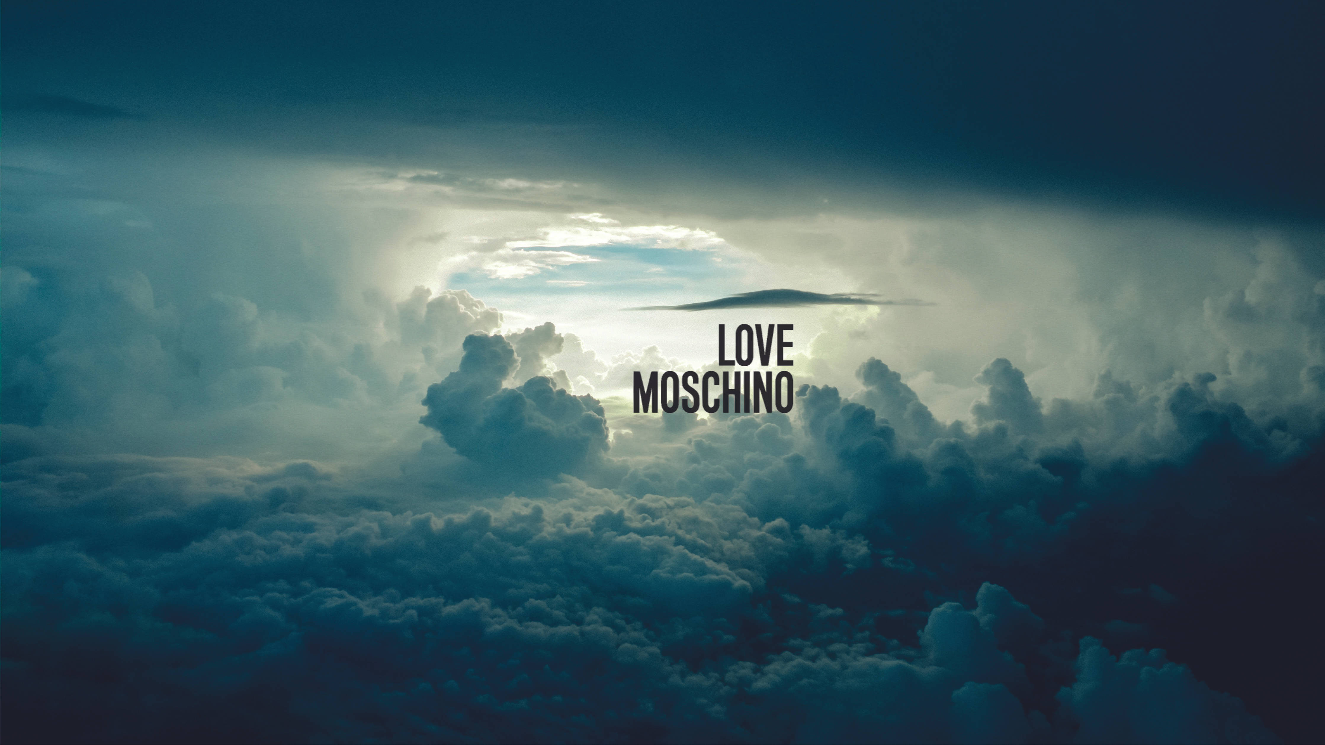 Love Moschino 雲層天空 壁紙 — 戲劇性的雲朵圍繞著明亮的太陽光芒旋轉，'LOVE MOSCHINO' 文字漂浮在藍灰色雲海之上。