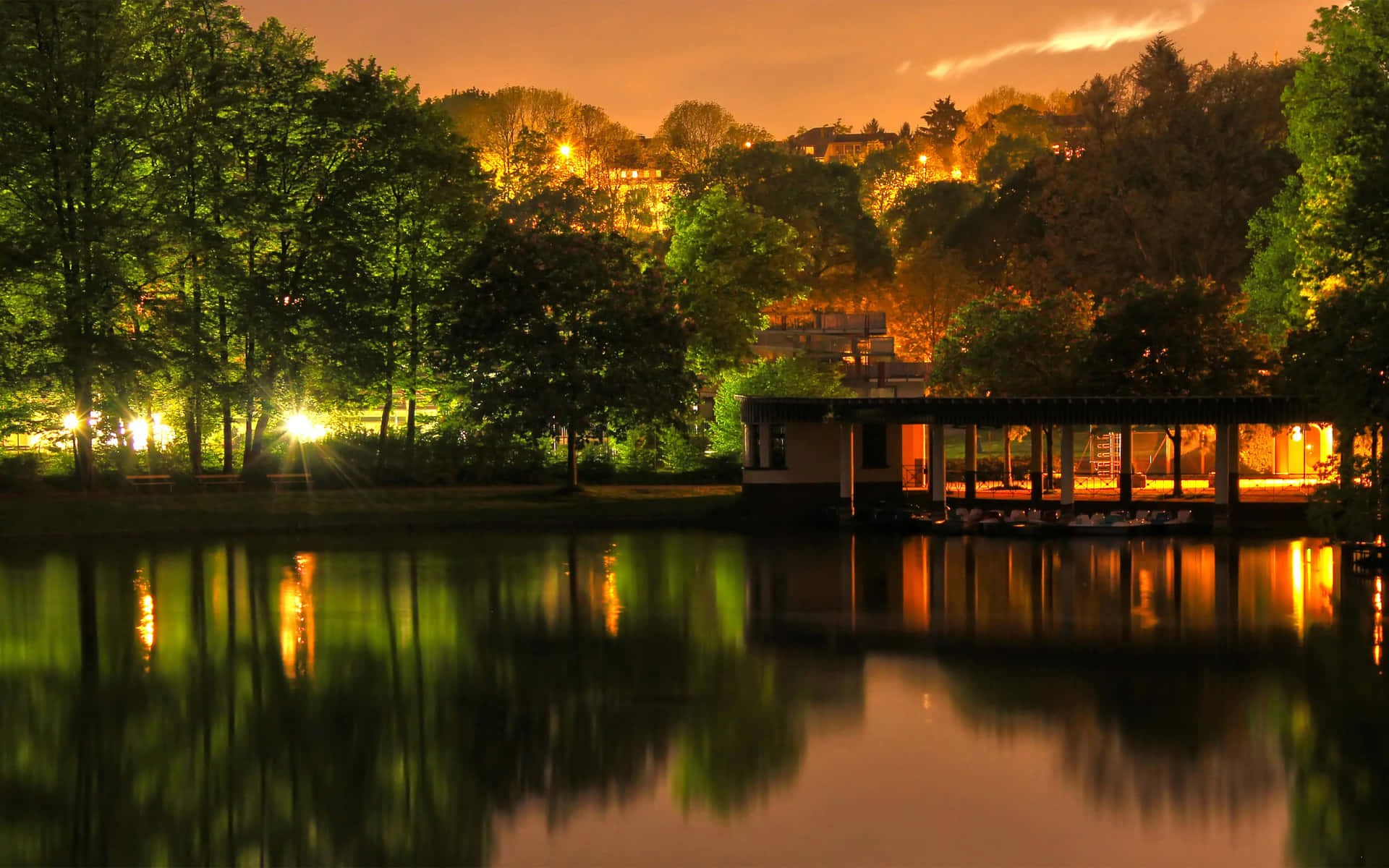  壁紙 — 寧靜的夜晚場景，湖邊建築物和樹木的溫暖燈光在橙色天空下美麗地倒映在平靜的水面上。