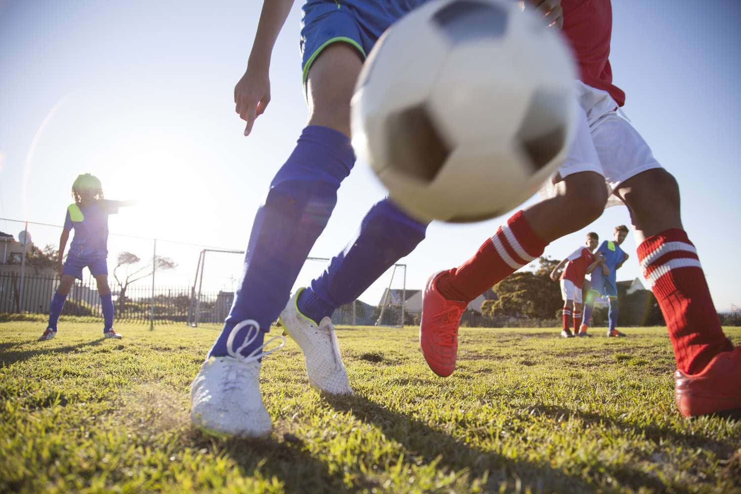 足球場——夢想成真的地方 壁紙 — 低角度拍攝足球運動員在陽光明媚的草地上比賽，前景有一個模糊的球。