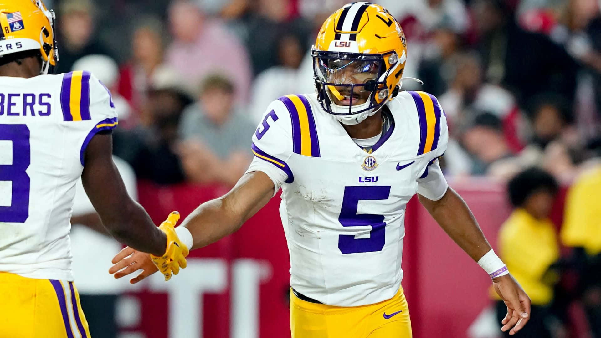 LSU四分衛與隊友擊掌慶祝 壁紙 — LSU 足球員身穿白色和金色球衣，在明亮的燈光下於球場上擊掌。
