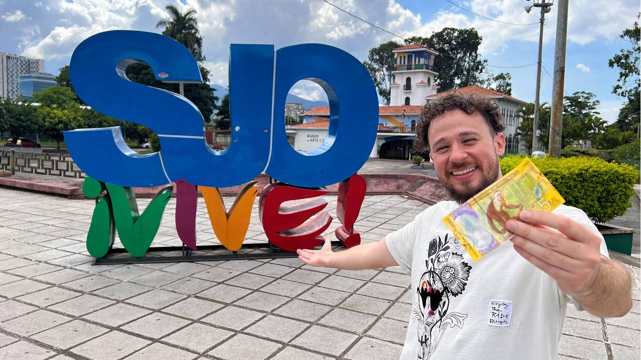 男人微笑並在色彩繽紛的「SUD VIVE！」地標附近展示鈔票。