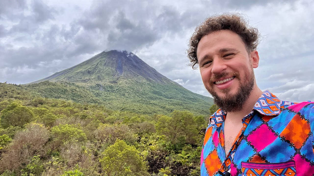 留鬍鬚和捲髮的男人在綠色火山前微笑。