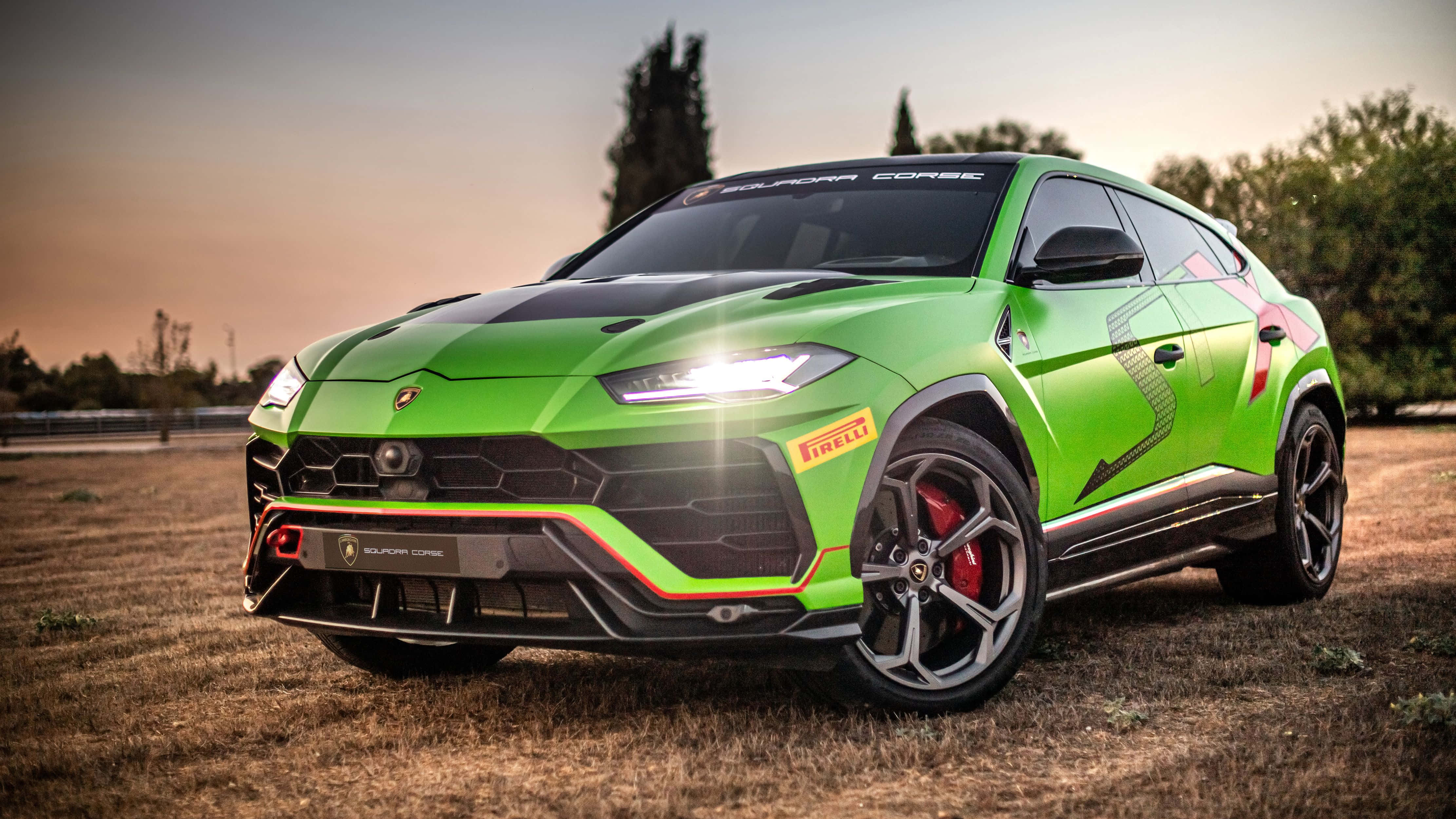 Lamborghini Urus 桌布 壁紙 — 一輛充滿活力的綠色林寶堅尼 Urus Performante 在日落時分停在乾草地上，車頭燈閃爍，紅色制動卡鉗清晰可見。