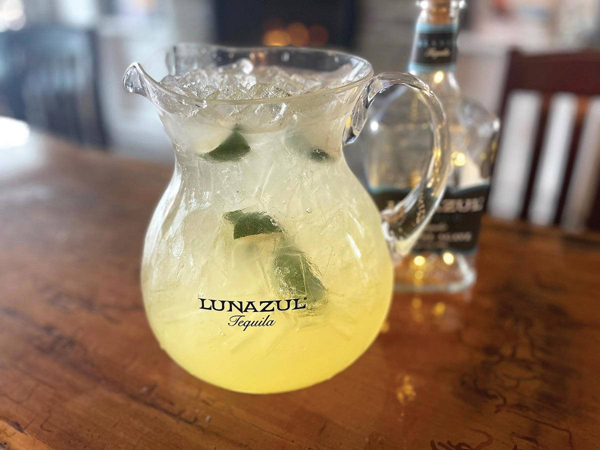 Lunazul 蒂克酒瑪格麗特調酒壺 壁紙 — 一個玻璃壺裝滿冰青檸角同淡黃色龍舌蘭雞尾酒，放喺木吧枱上，背景有支酒。