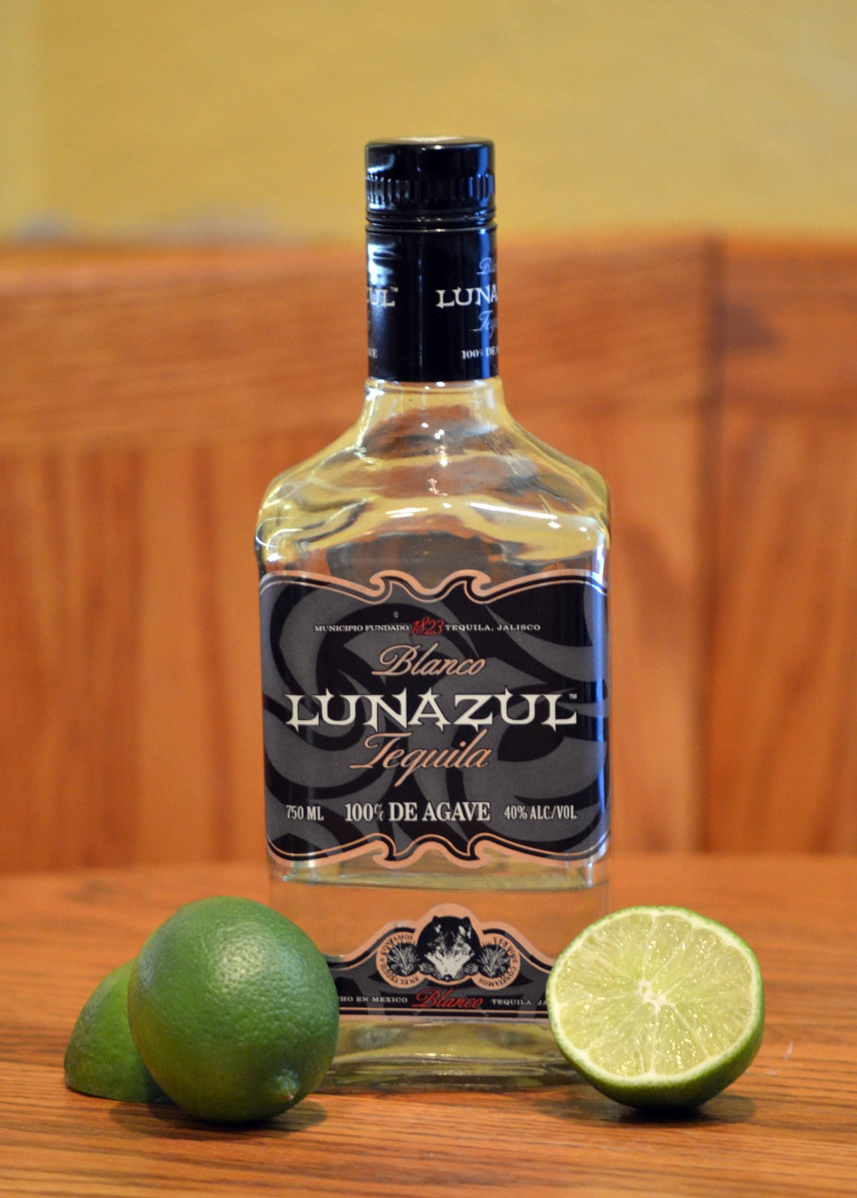 品嚐Lunazul Blanco龍舌蘭酒與新鮮青檸 壁紙 — 一個透明的Lunazul Blanco龍舌蘭酒瓶放在木桌上，旁邊有完整和切半的青檸。