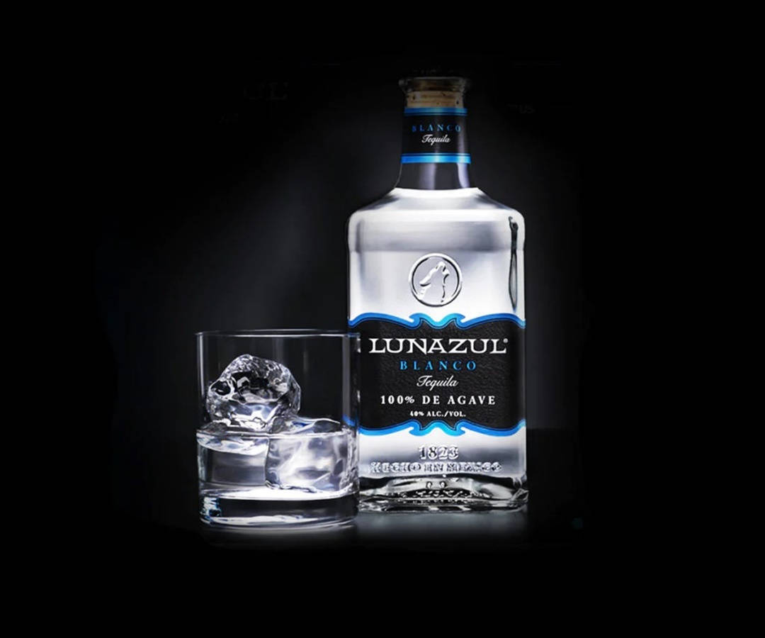 Lunazul Blanco 蒂克酒玻璃杯 壁紙 — 一瓶流線型的Lunazul Blanco龍舌蘭酒放在一個裝有大冰塊的玻璃杯旁邊，背景為深色。