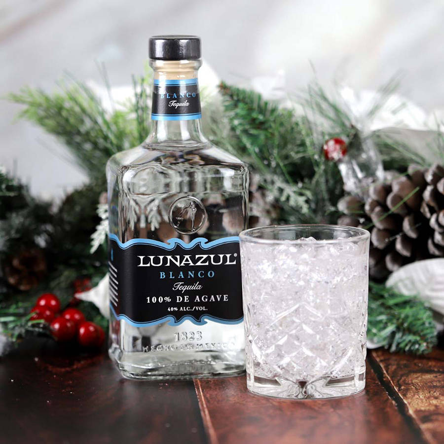 Lunazul Blanco龍舌蘭酒聖誕裝飾 壁紙 — 一瓶Lunazul Blanco龍舌蘭酒和一杯冰塊放在木桌上，旁邊有節日松枝和紅莓。