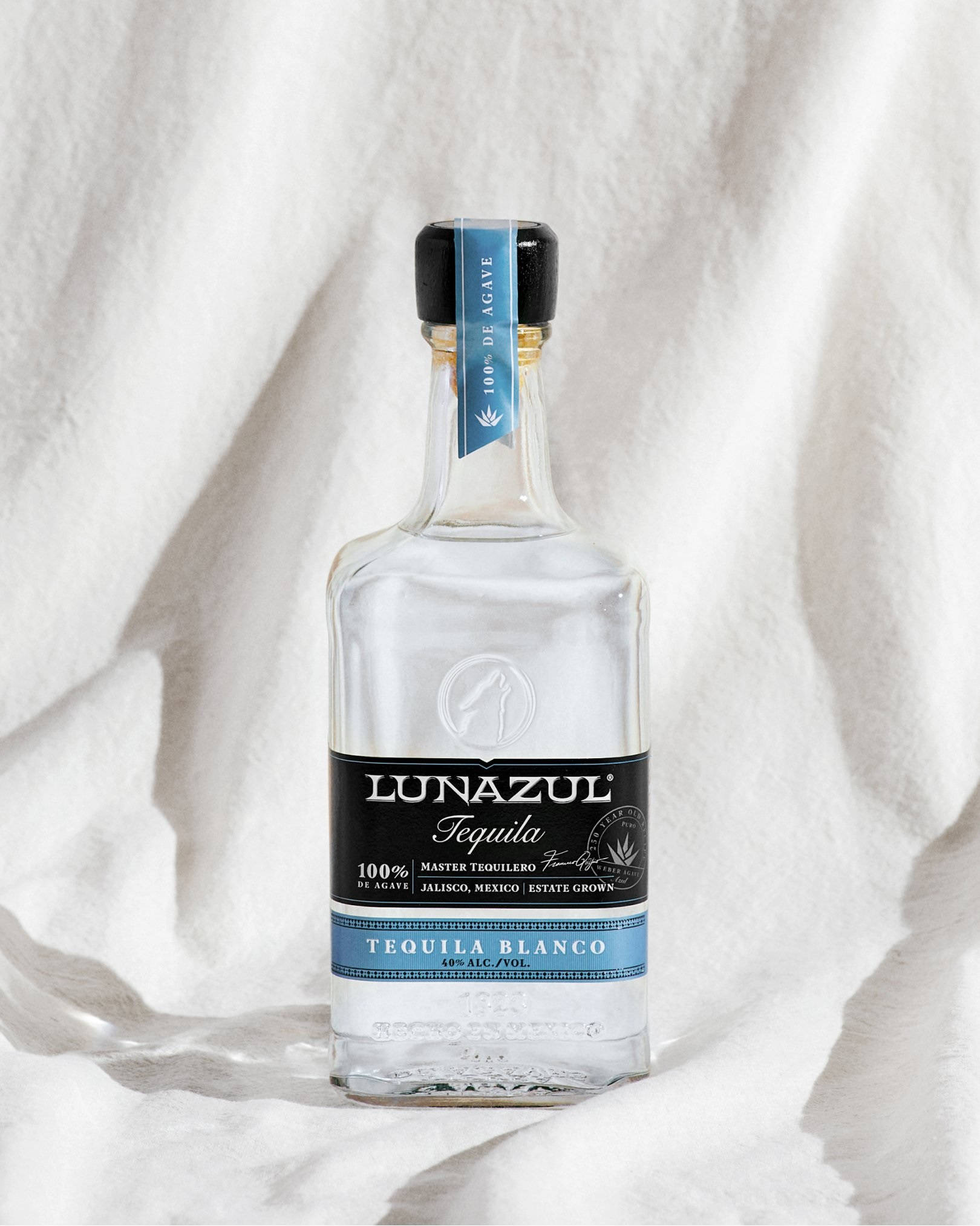 Lunazul Blanco龍舌蘭酒白色布料 壁紙 — 一支透明的Lunazul Tequila Blanco酒瓶放在柔軟的白色布料上，在明亮的自然光下。