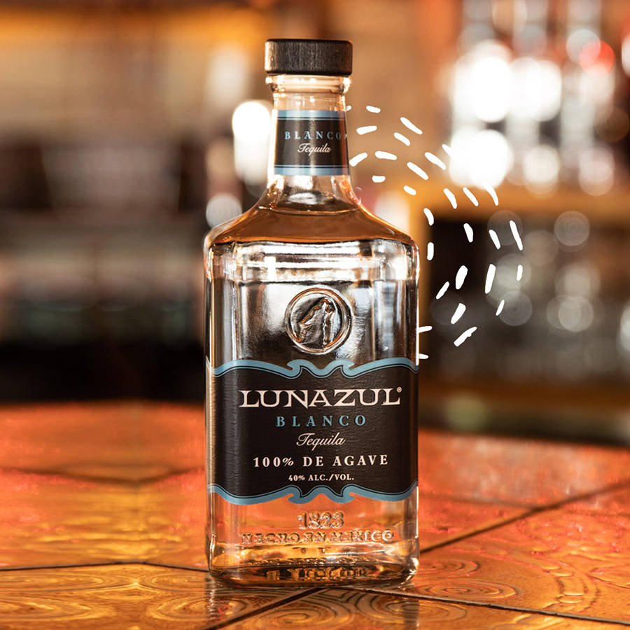Lunazul Blanco龍舌蘭酒白色圖案效果 壁紙 — 一個透明的Lunazul Blanco龍舌蘭酒瓶放在溫暖的木吧檯上，背景有模糊的玻璃器皿和金色燈光。