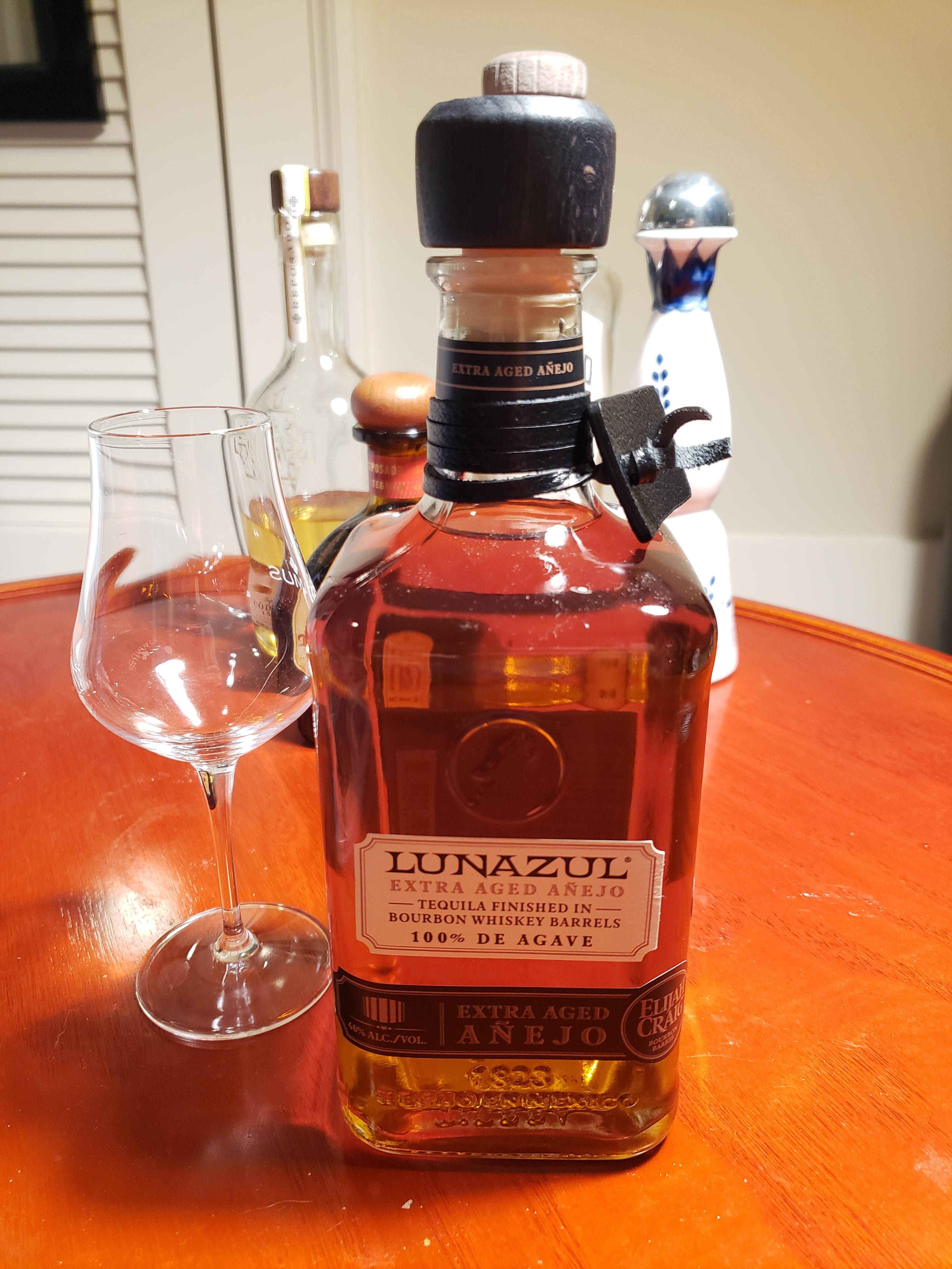 Lunazul Extra Aged Anejo - 頂級藍色龍舌蘭酒 壁紙 — 一瓶Lunazul Extra Aged Añejo龍舌蘭酒放在溫暖的木桌上，旁邊有一個空的品酒杯。