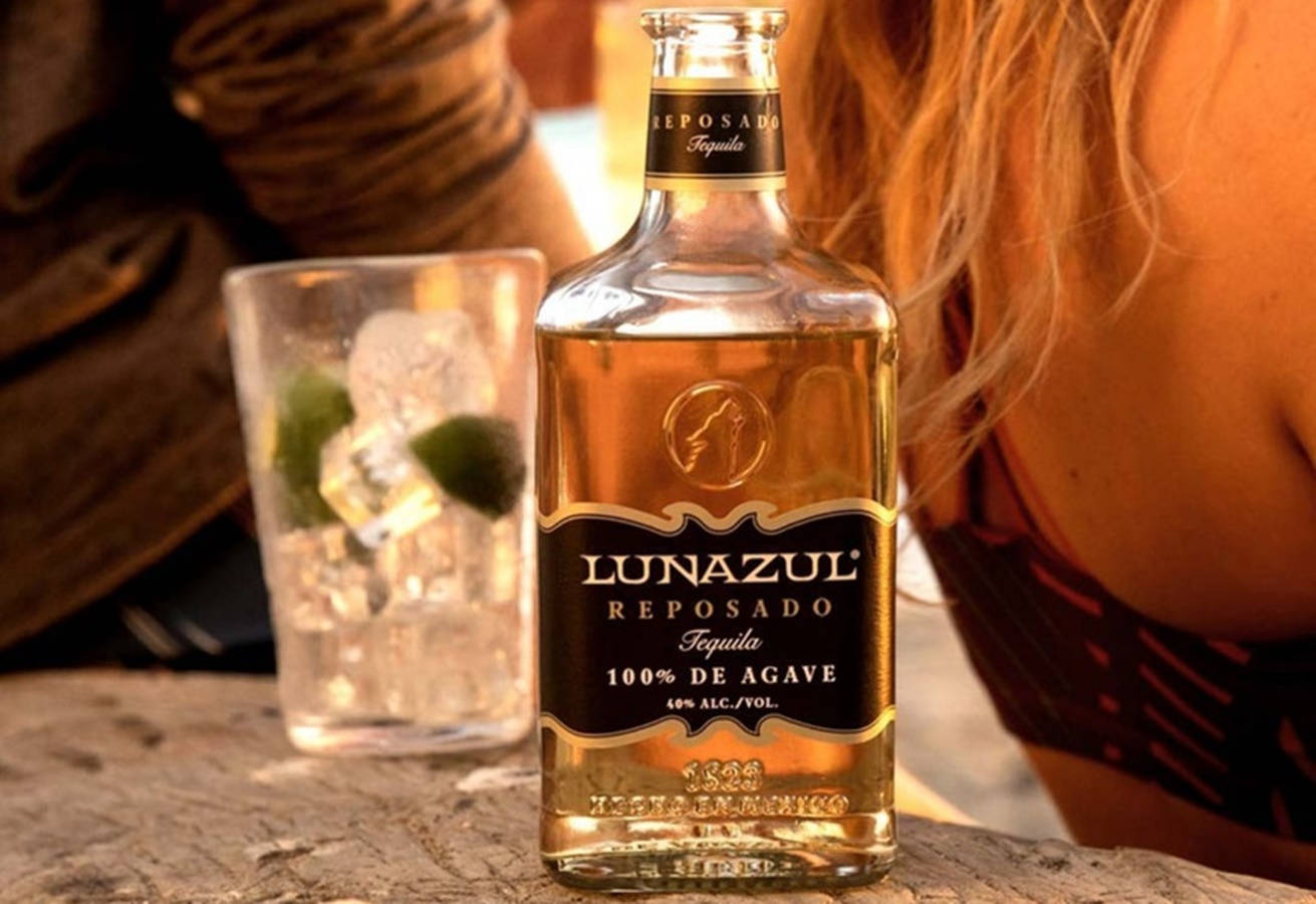 Lunazul Reposado 冰杯 壁紙 — 一瓶Lunazul Reposado龍舌蘭酒放在石桌上，旁邊有一杯加冰和青檸的酒杯，一位女士靠在附近。