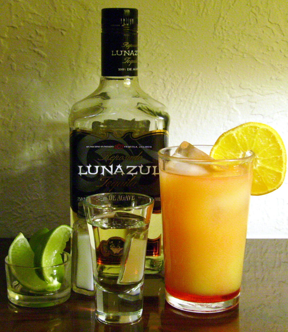 品味風味，Lunazul Reposado 蒸餾龍舌蘭酒 壁紙 — 一瓶Lunazul龍舌蘭酒放在一個高身橙色雞尾酒杯旁邊，杯中有冰塊和青檸裝飾，背景是紋理牆壁。