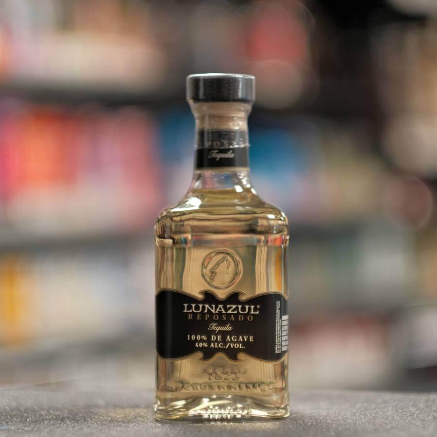 Lunazul Reposado龍舌蘭酒淡黃色 壁紙 — 一個透明的Lunazul Reposado龍舌蘭酒瓶放在灰色表面上，背景是模糊的商店貨架。